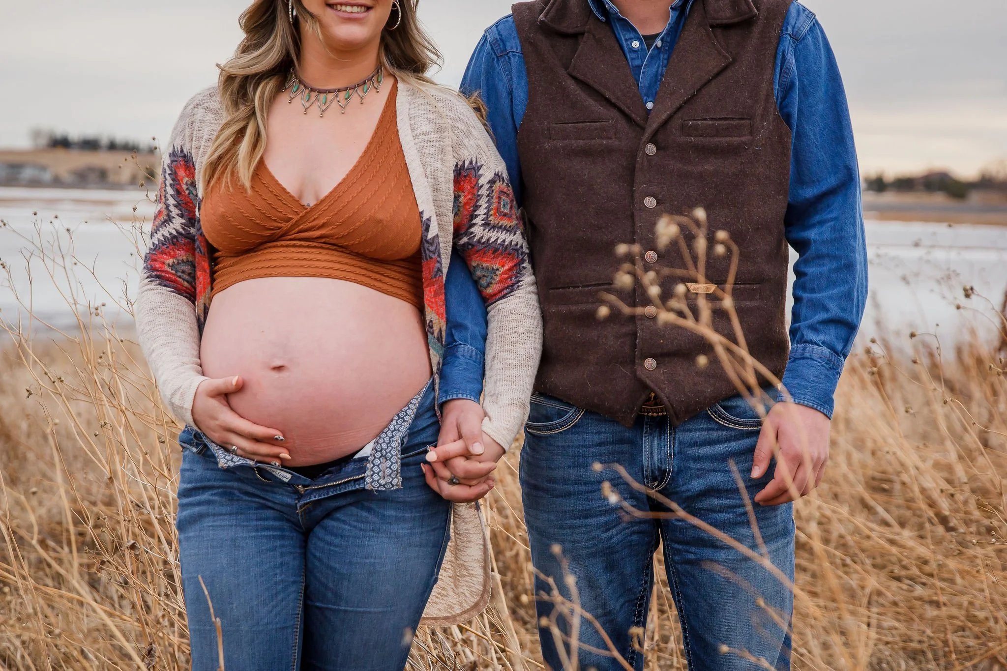 A pregnant woman and a man holding hands outdoors in a field of tall dried grass, with water and a distant shoreline in the background during a cloudy day. Maternity photography session by Ralph Klein park in Calgary, Alberta. 