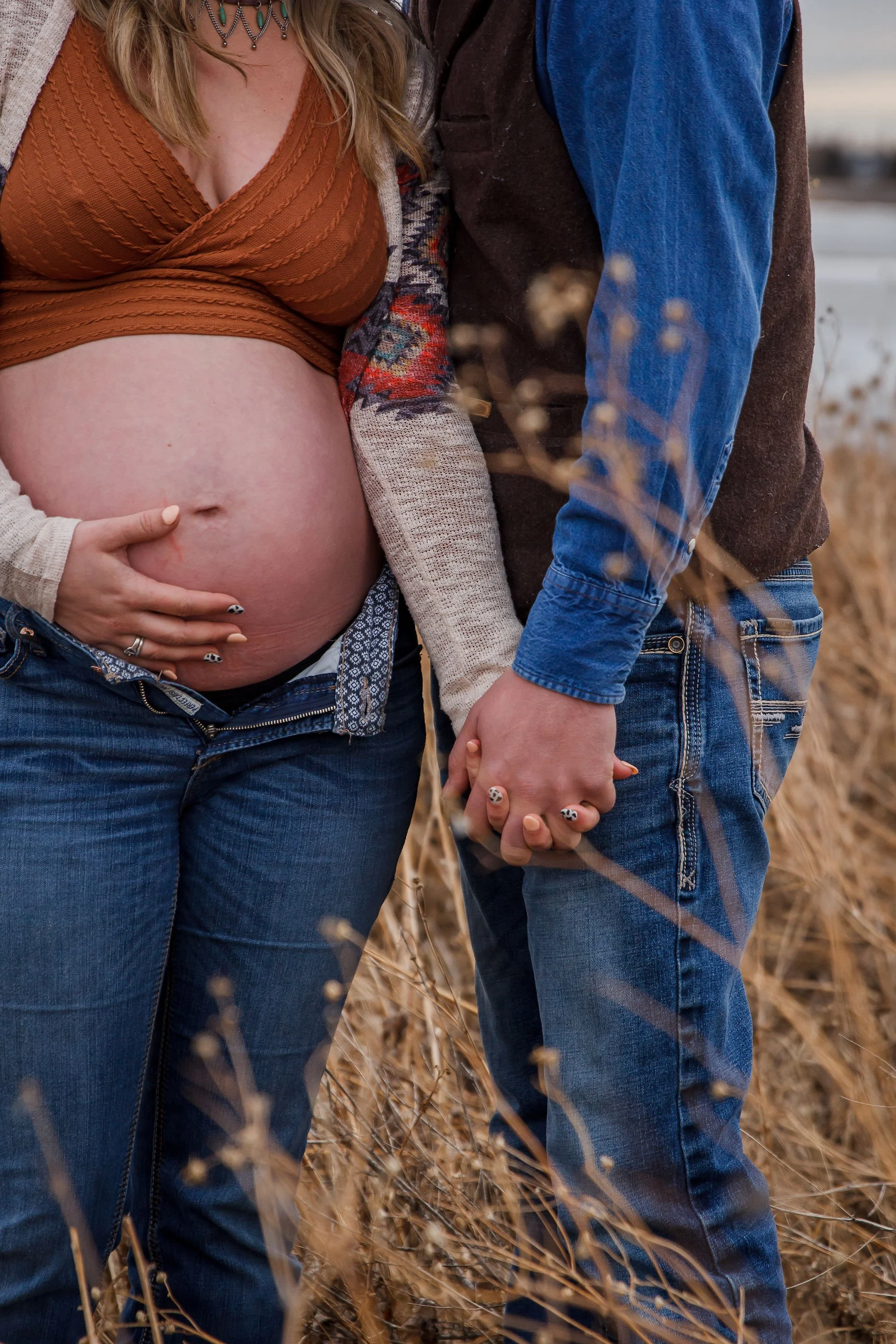 A pregnant woman and a man holding hands outdoors in a field of dry grass, with the woman's hand resting on her baby bump. Maternity photography session by Ralph Klein park in Calgary, Alberta. 