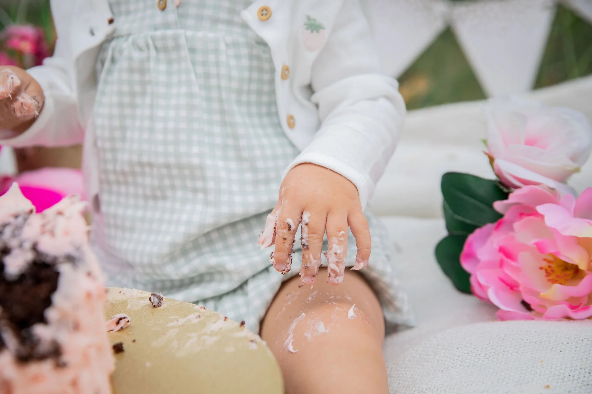 Child's hand covered in cake and frosting, with a birthday cake and pink flowers nearby. Outdoor cake smash photos at the Inglewood Bird Sanctuary in Calgary, Alberta. 