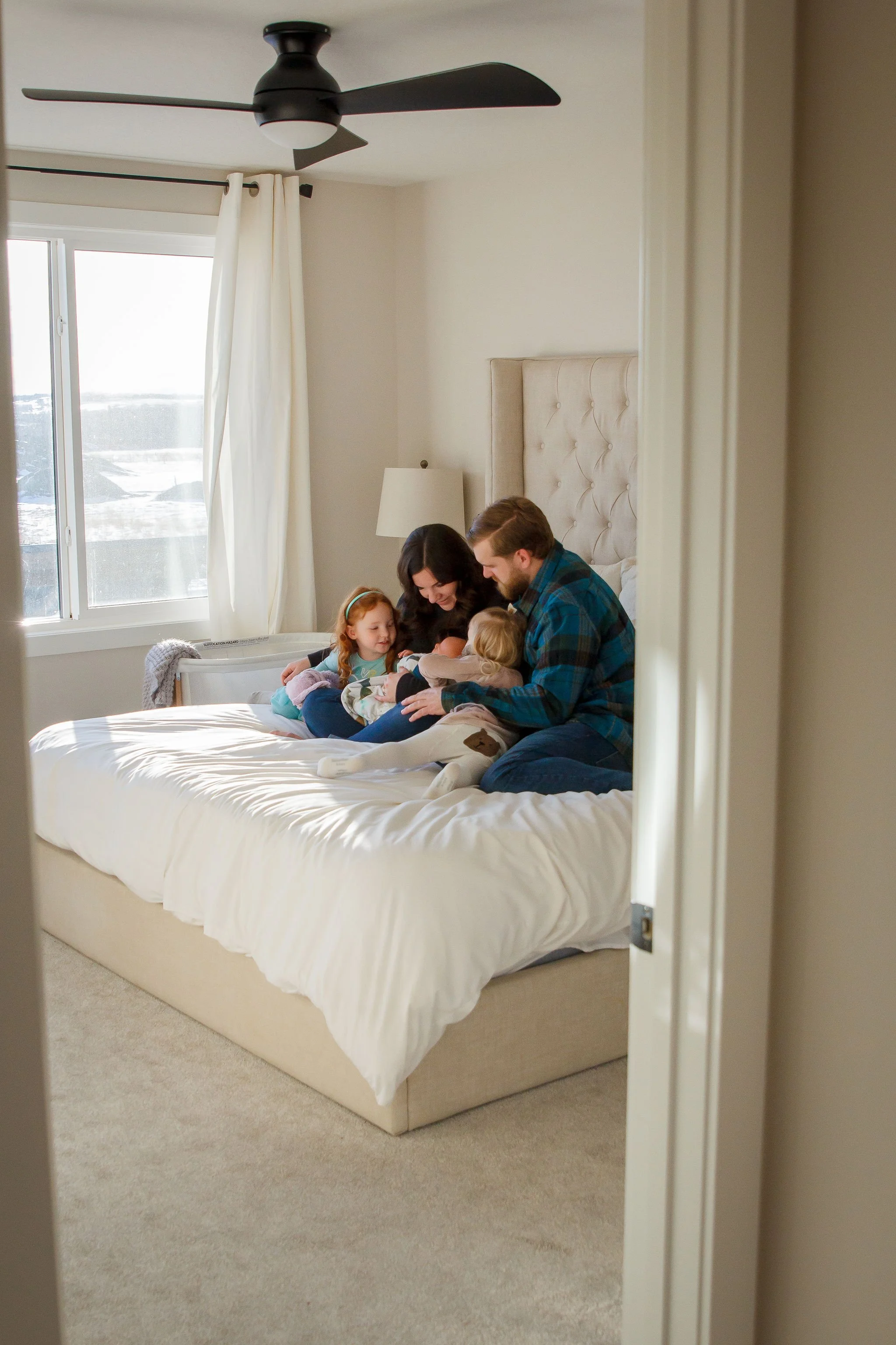 Family of five sitting on bed, bonding with two young children, in a brightly lit bedroom with white walls, beige headboard, window with curtains, and ceiling fan.