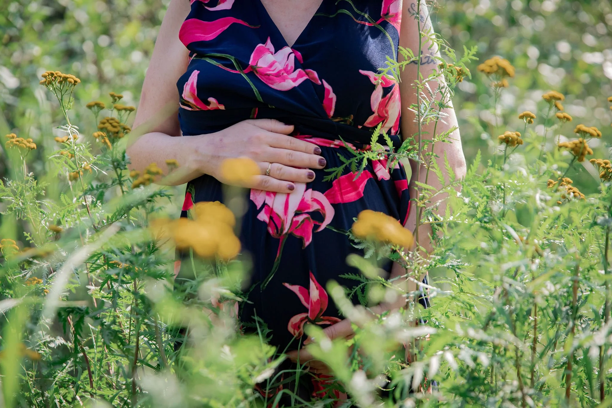 A woman in a dark floral dress holding her pregnant belly while standing in a field of yellow wildflowers. maternity family photography at Bow Valley Habitat Station in Calgary Alberta