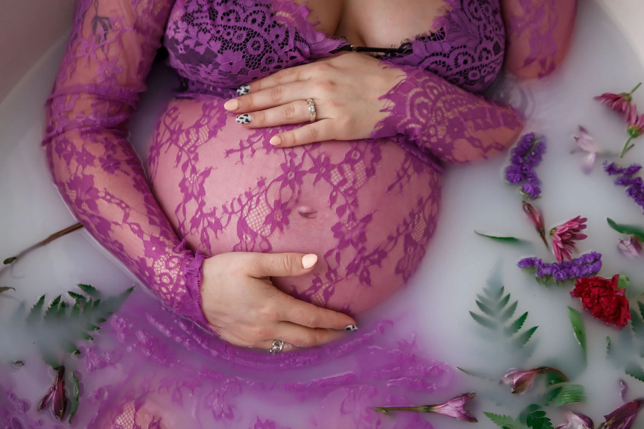 A pregnant woman wearing a purple lace dress, lying in milk with various colored flowers floating around her, gently holding her belly with both hands.