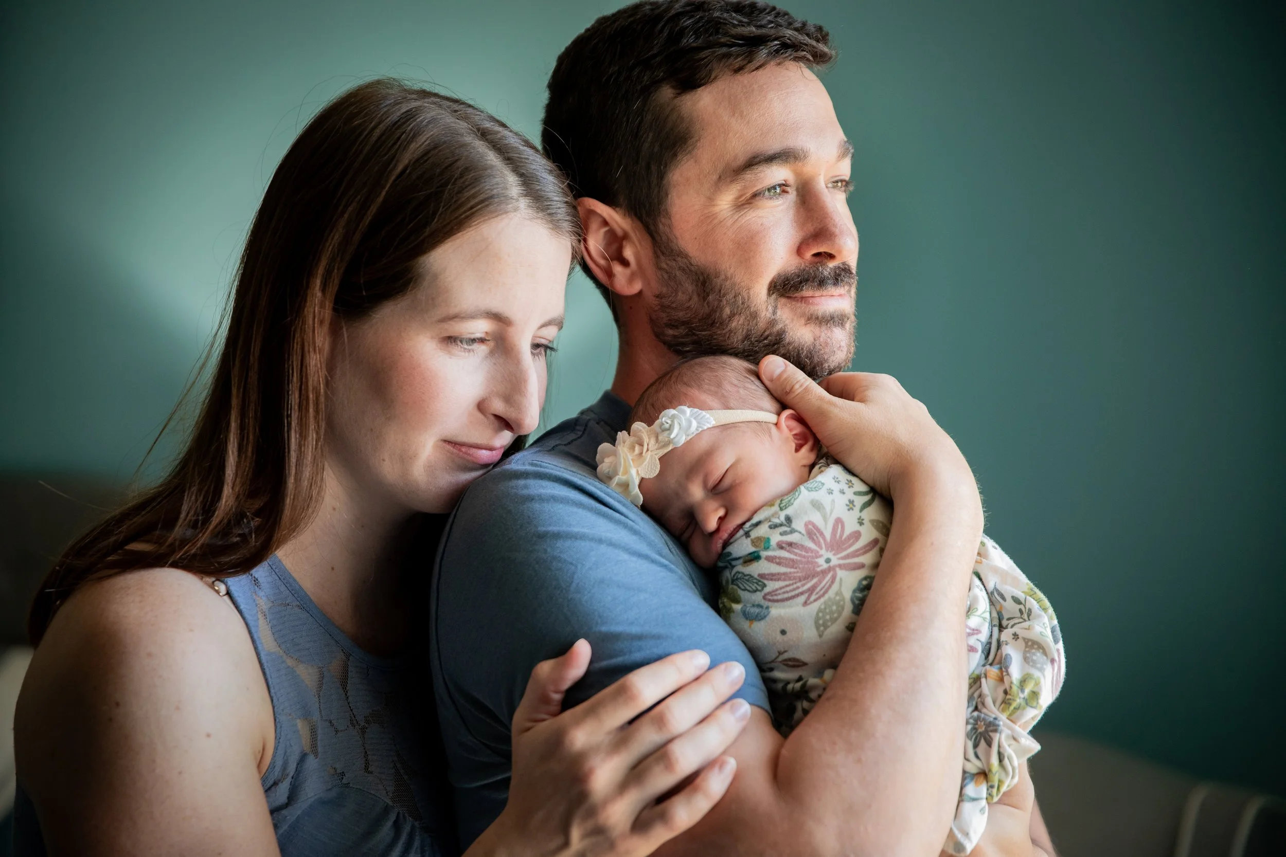 A family of three with a woman, a man, and a sleeping newborn baby wrapped in a floral blanket. The woman and man are close together, gently cradling the baby, with peaceful and content expressions in this in home newborn session. Calgary, Alberta. 