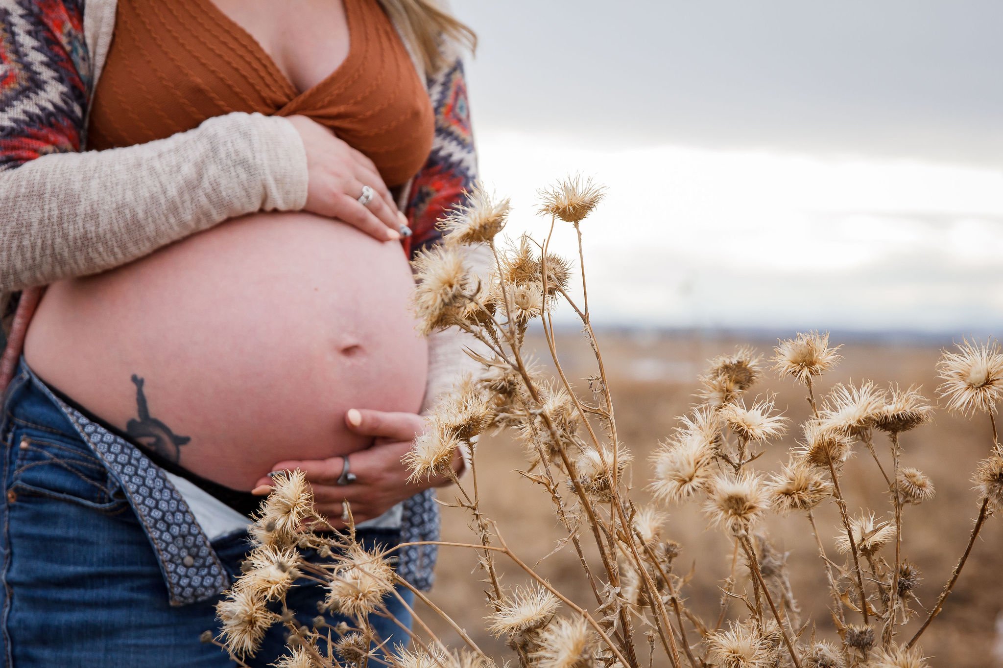 A pregnant woman is standing outdoors in a field with dried plants, holding her belly with one hand and the other hand resting on her stomach. Maternity photography session by Ralph Klein park in Calgary, Alberta. 