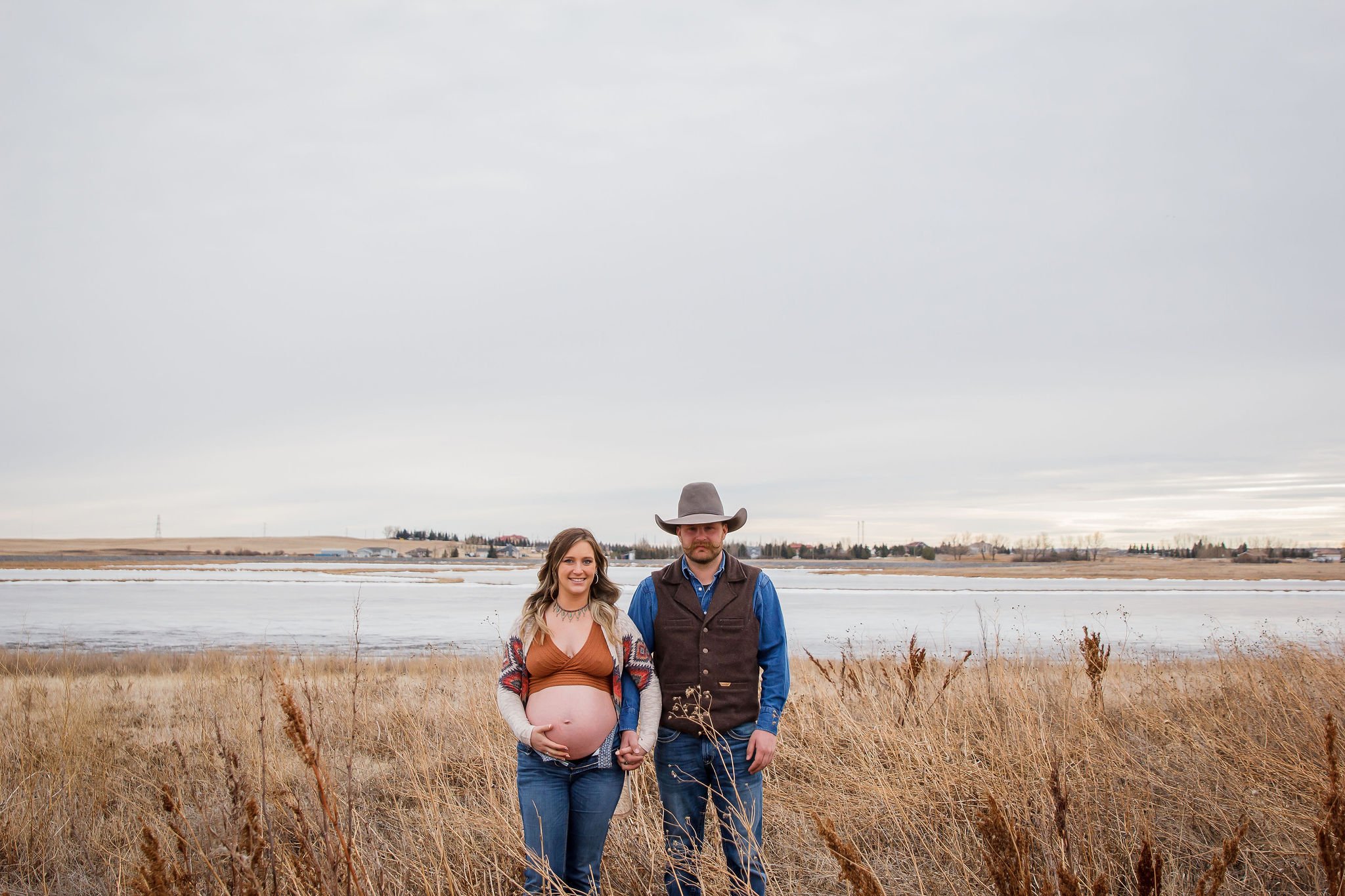 A pregnant woman and a man holding hands standing in a field near a body of water with dry grass and overcast sky. Maternity photography session by Ralph Klein park in Calgary, Alberta. 