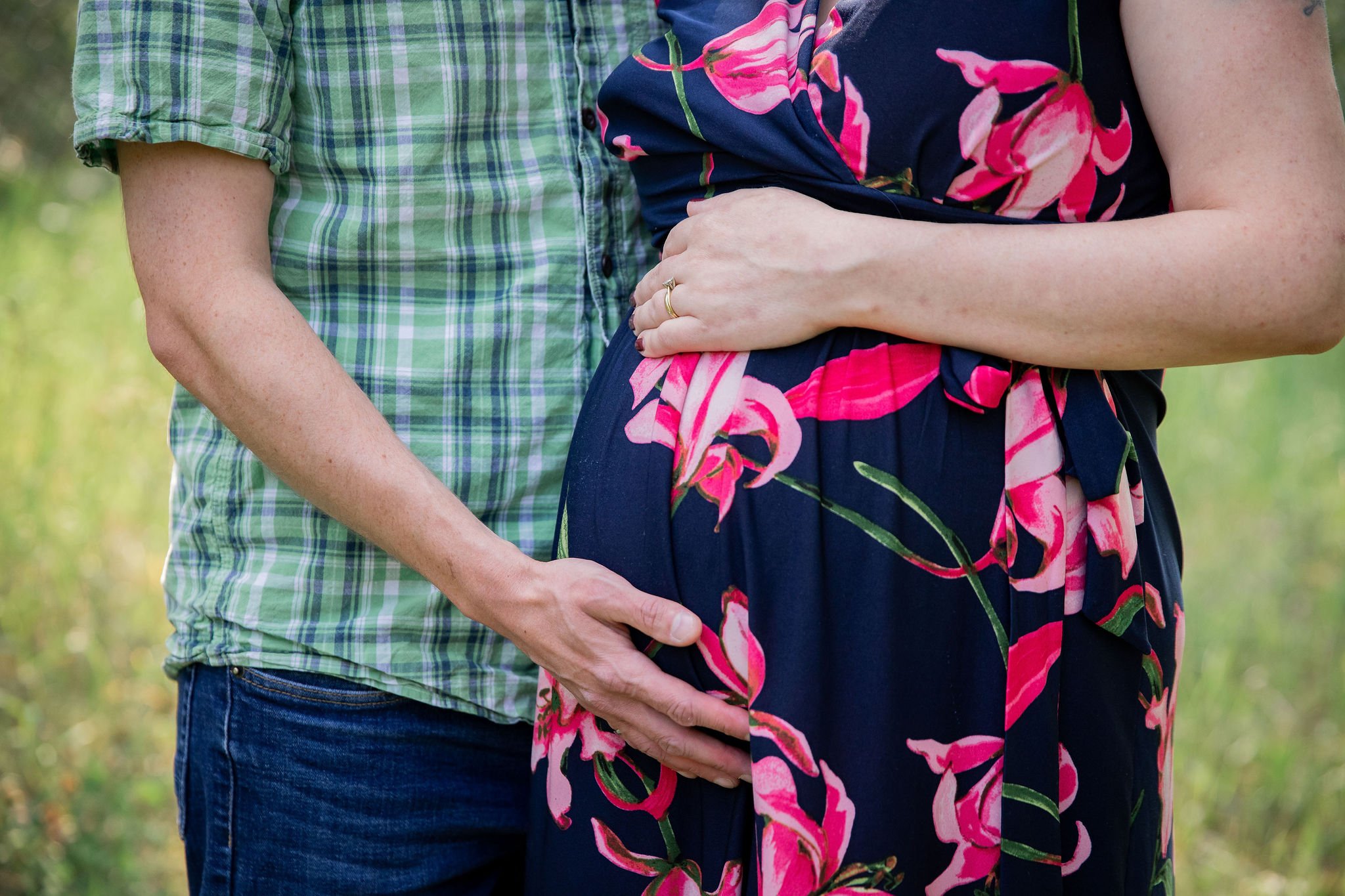 A pregnant woman wearing a floral dress and a man in a checkered shirt are holding her belly and waist outdoors. maternity family photography at Bow Valley Habitat Station in Calgary Alberta