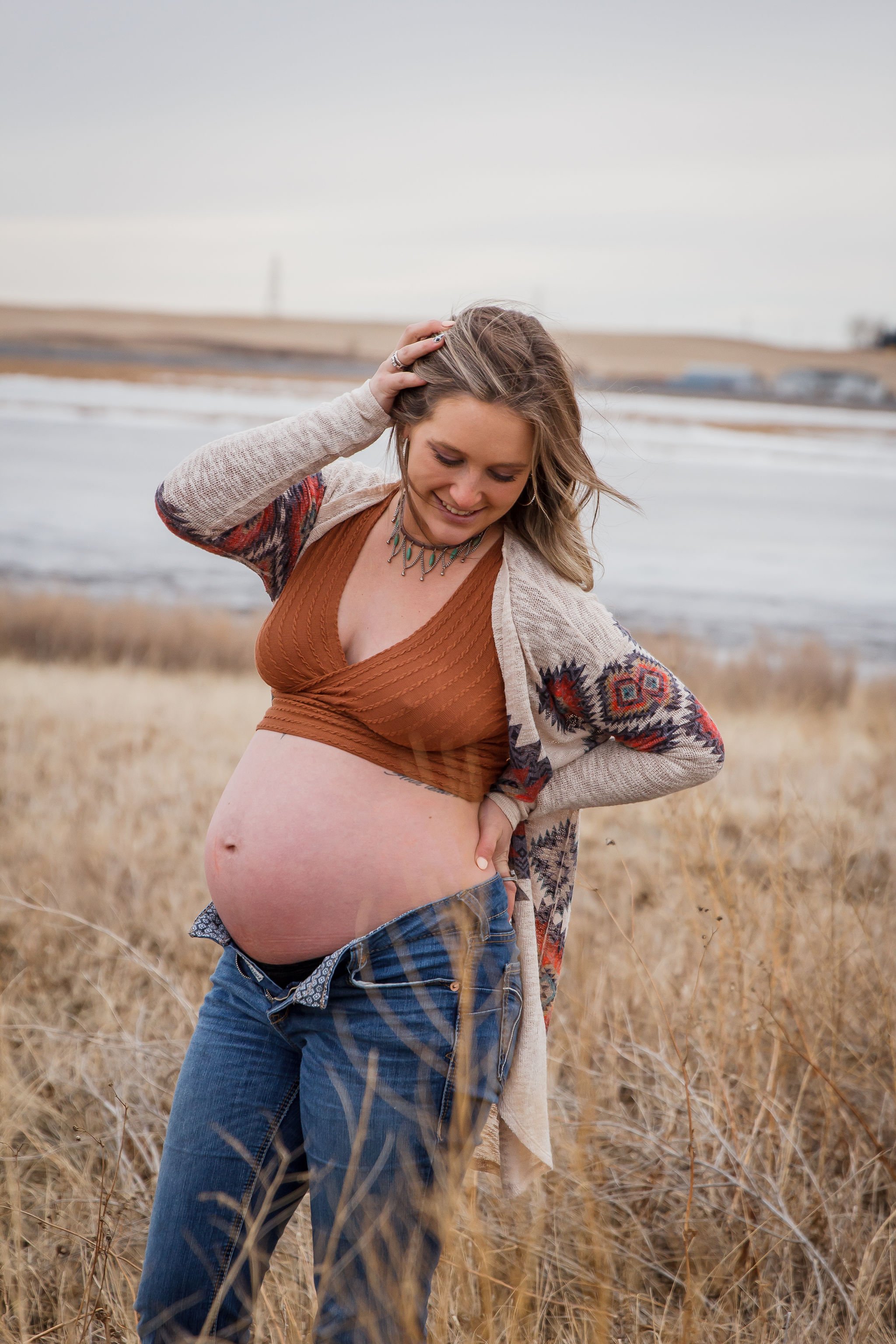 Pregnant woman wearing a brown top and a patterned jacket standing in a field of dry grass near water, smiling and touching her hair. Maternity photography session by Ralph Klein park in Calgary, Alberta. 