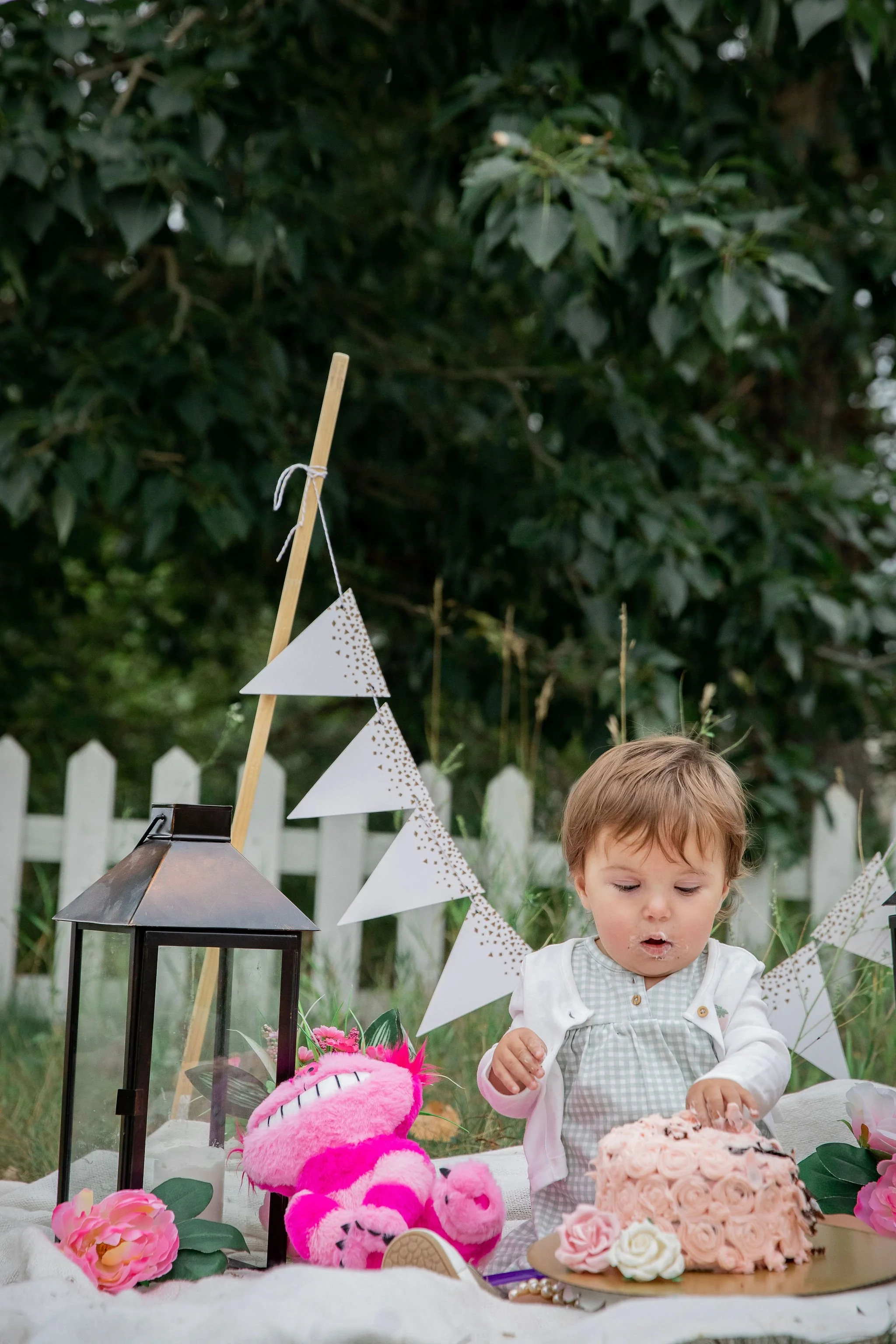 Child celebrating birthday outdoors with pink cake, stuffed animal, lantern, and bunting flags, in front of a white picket fence. Outdoor cake smash photos at the Inglewood Bird Sanctuary in Calgary, Alberta. 