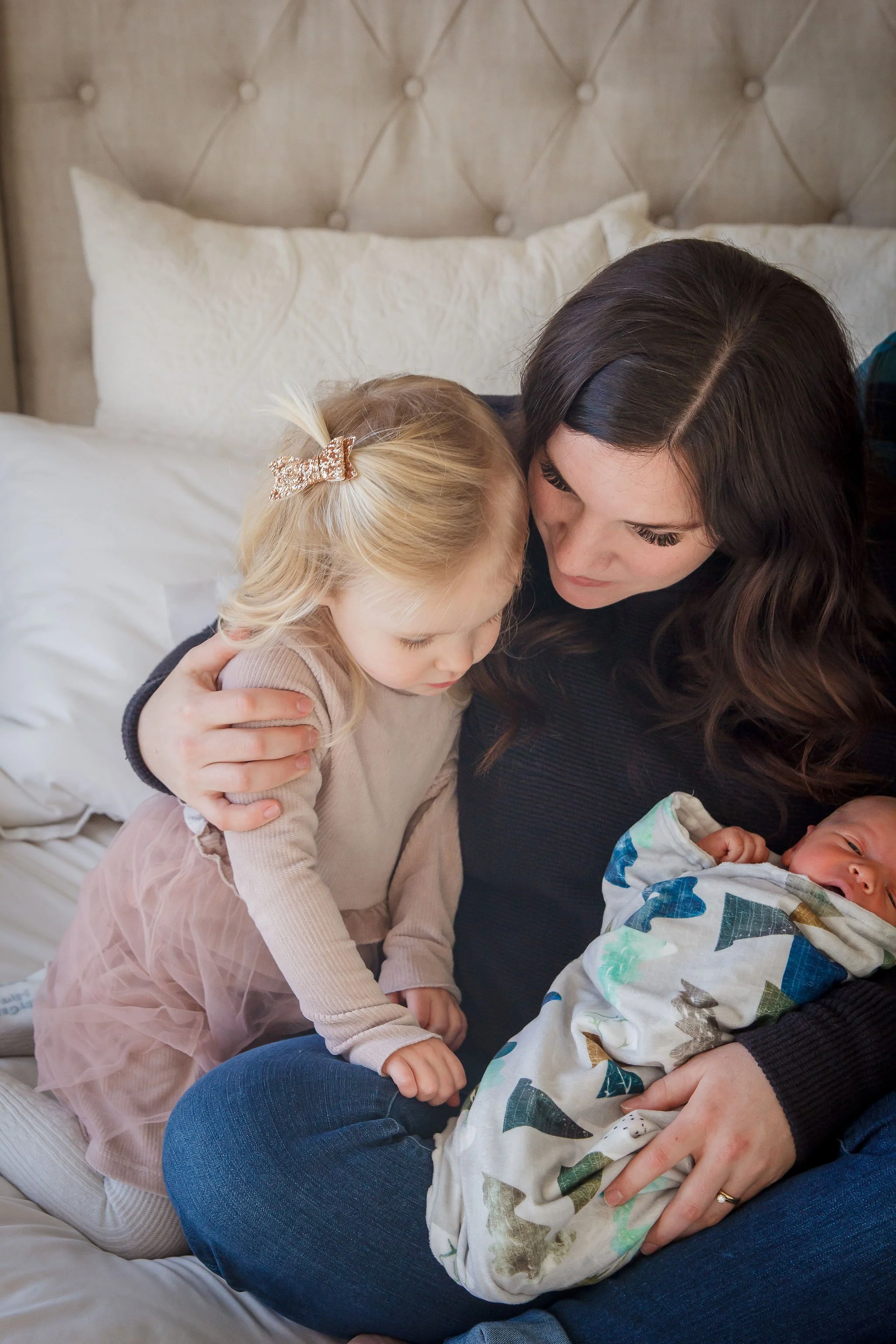 A woman sitting on a bed with a young girl and a newborn baby, holding the baby in her arms while the girl looks on.