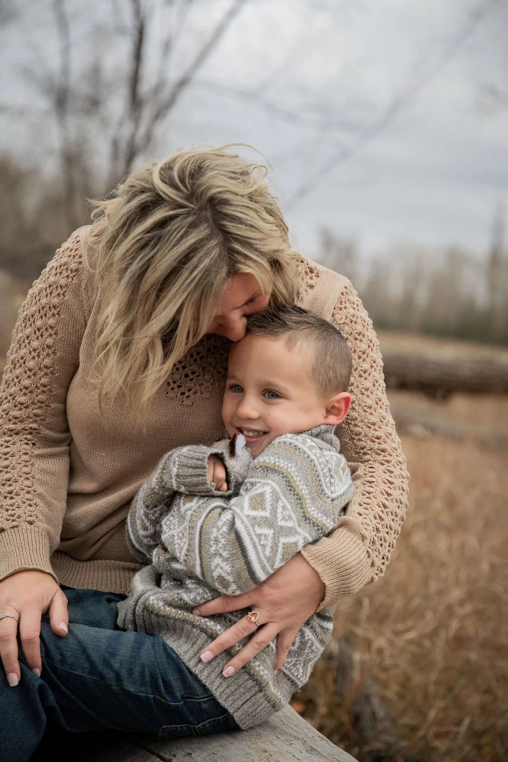 A woman with blonde hair hugging a young boy with short hair, outdoors in a field with bare trees and overcast sky in Calgary, Alberta. 