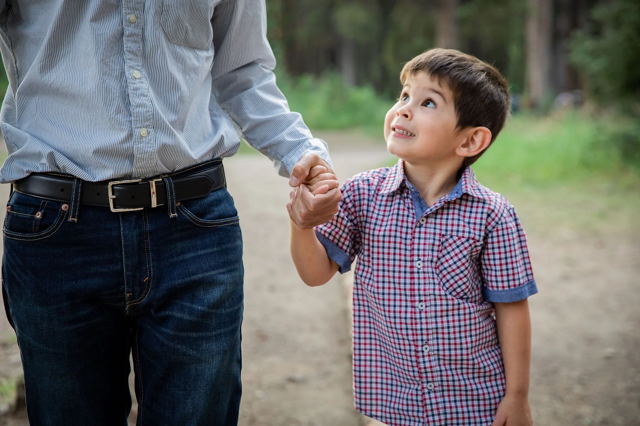A young boy holding hands with an adult, looking up with a smile, outdoors in a wooded area. Bebo Grove, Calgary Alberta. 