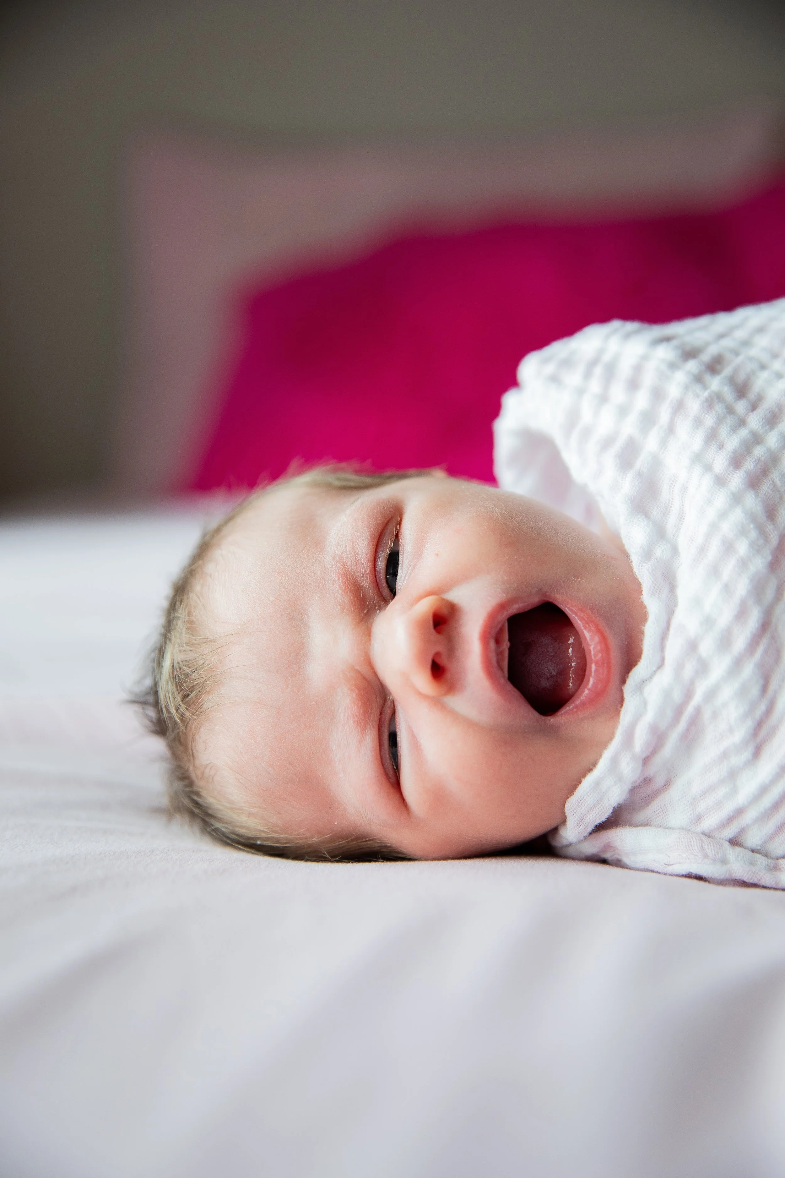 Yawning baby lying on a bed with a blurred pink and red background. In home newborn photography in Calgary, Alberta. 