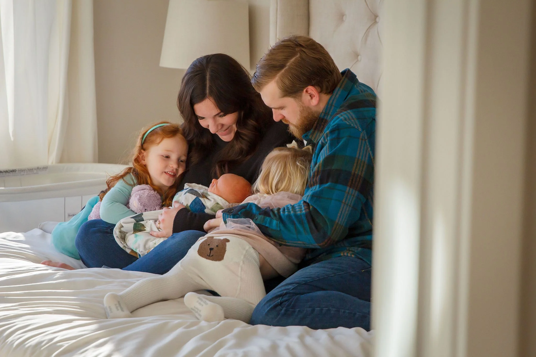 A family gathers on a bed, with a woman and a man holding infants, while a young girl and a toddler watch and smile.