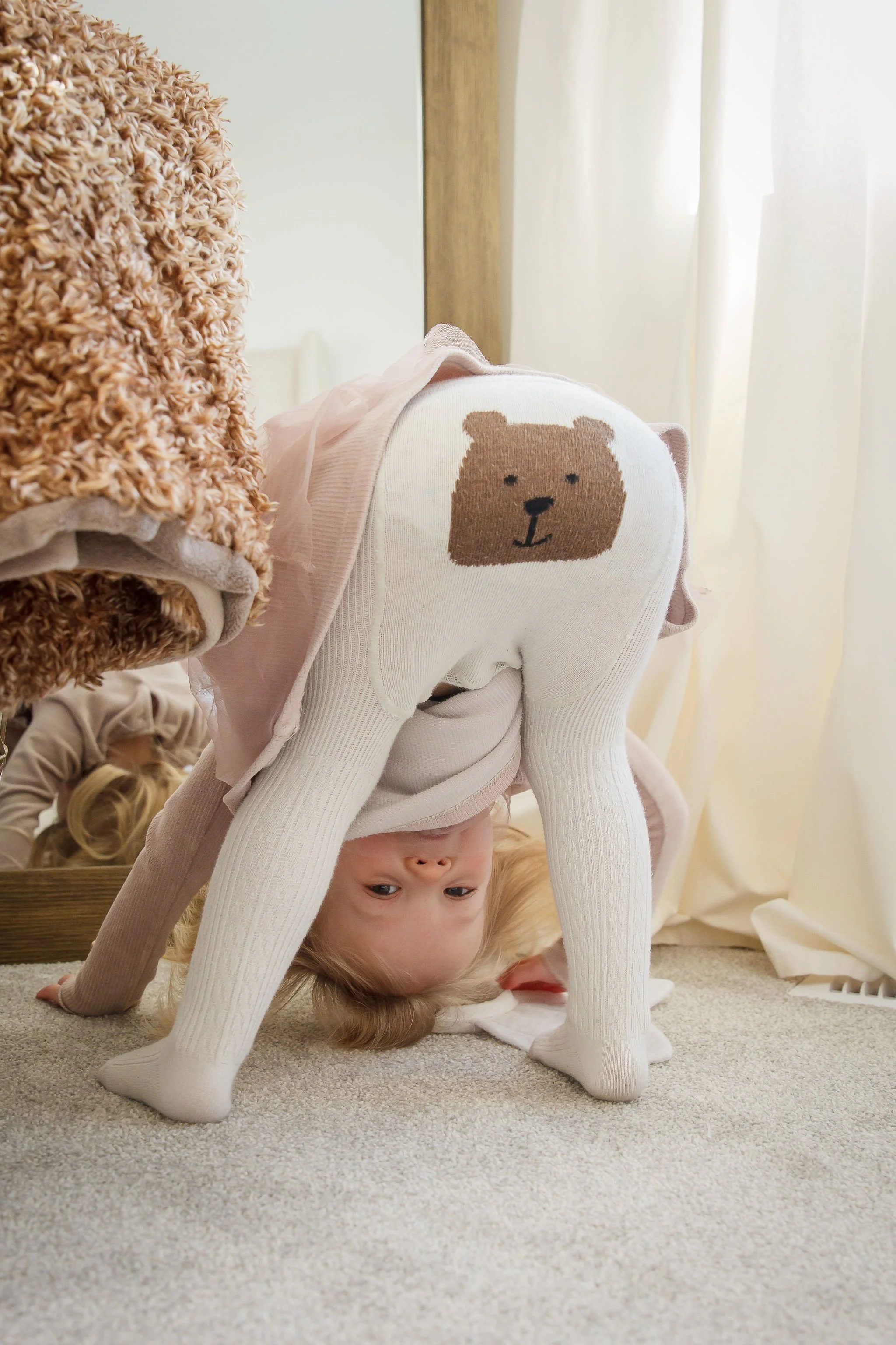Child playing with their head upside down, wearing a white sweater with bear face on the back, in a room with carpet and curtains.
