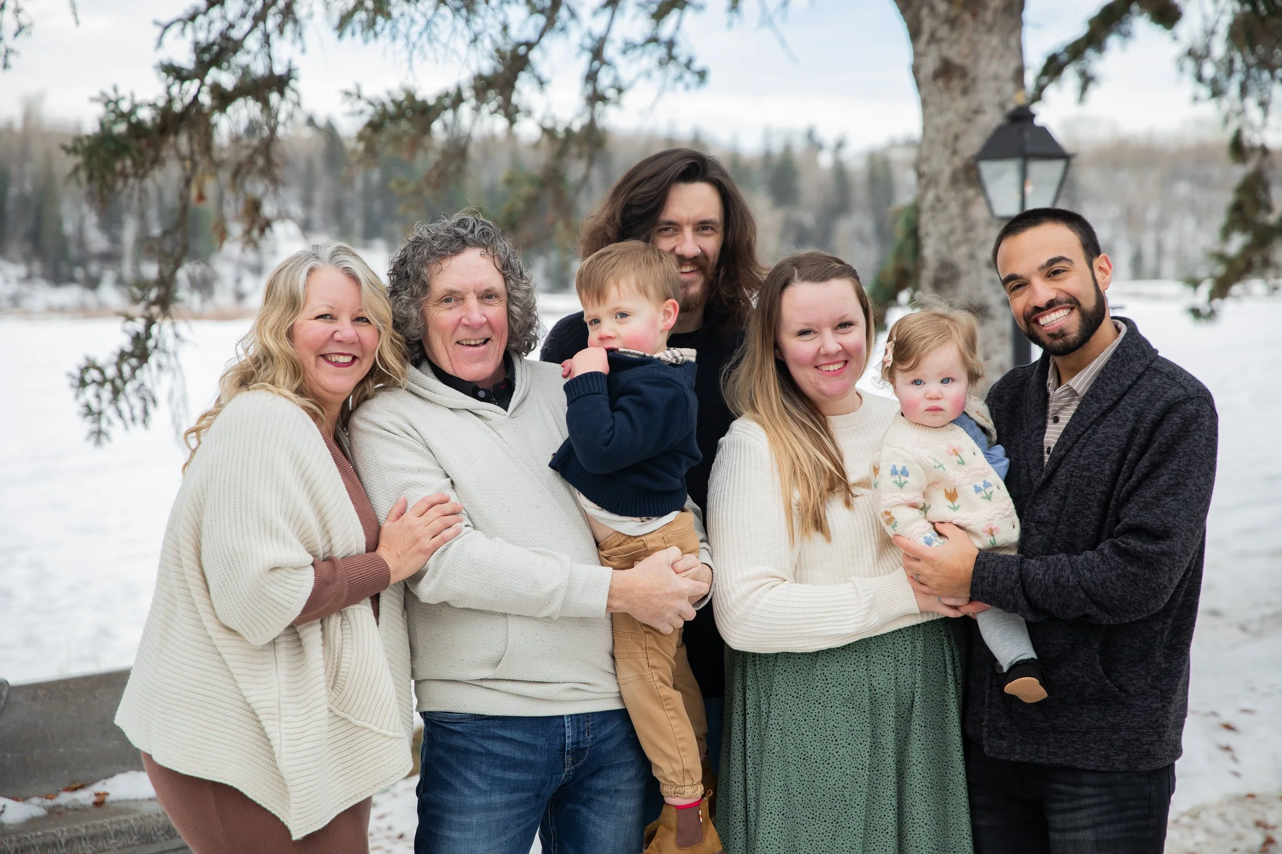 A diverse family of seven stands outdoors in a snowy park during winter, smiling at the camera with a lake and snow-covered trees in the background in 
Calgary, Alberta. 
