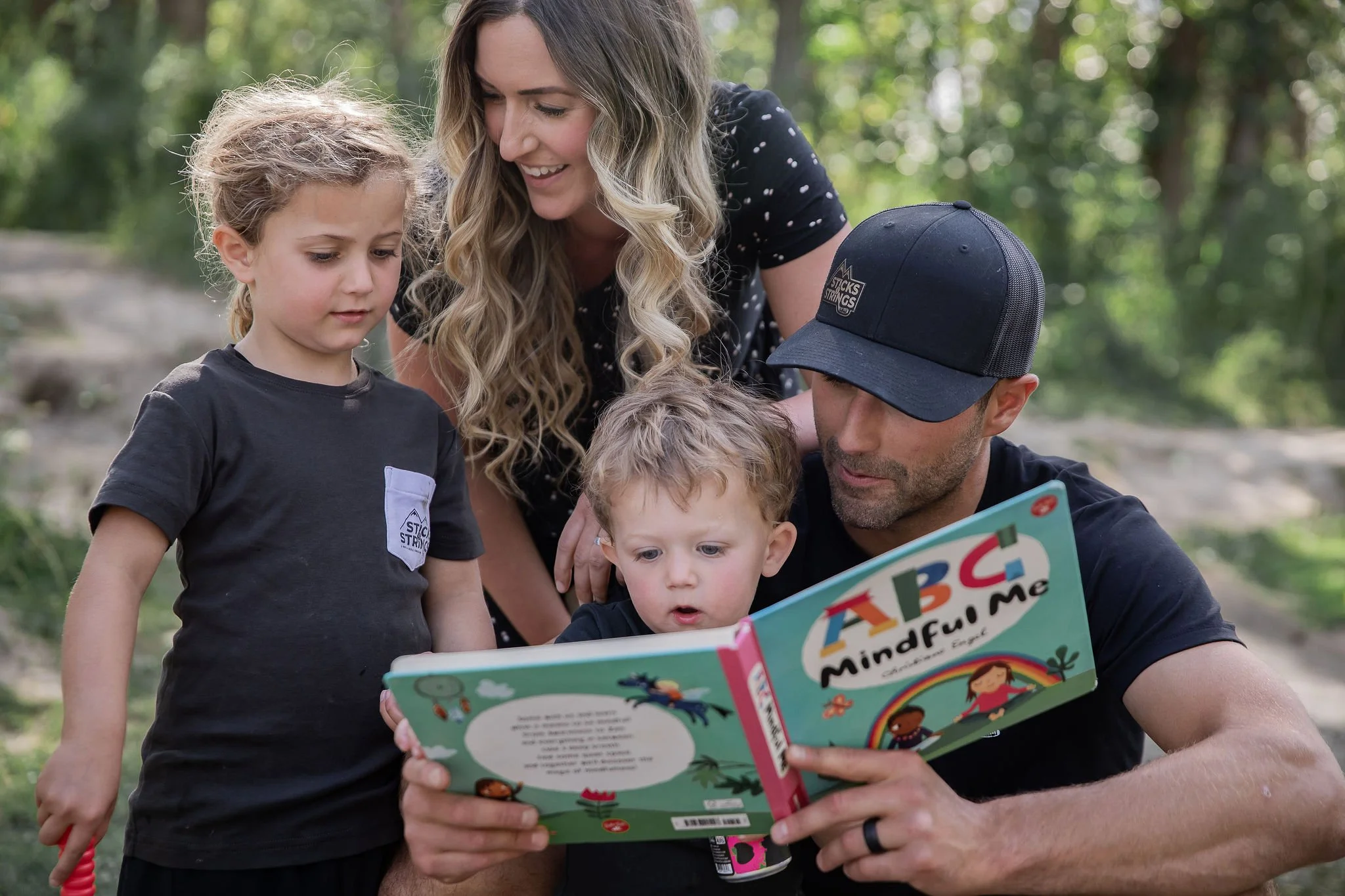 A family of four sitting outdoors with the father reading a colorful children's book titled 'ABC Mindful Me' to his two young children, while the mother leans over to look at the book. Branding session with Lauren and Jordan Eberle.