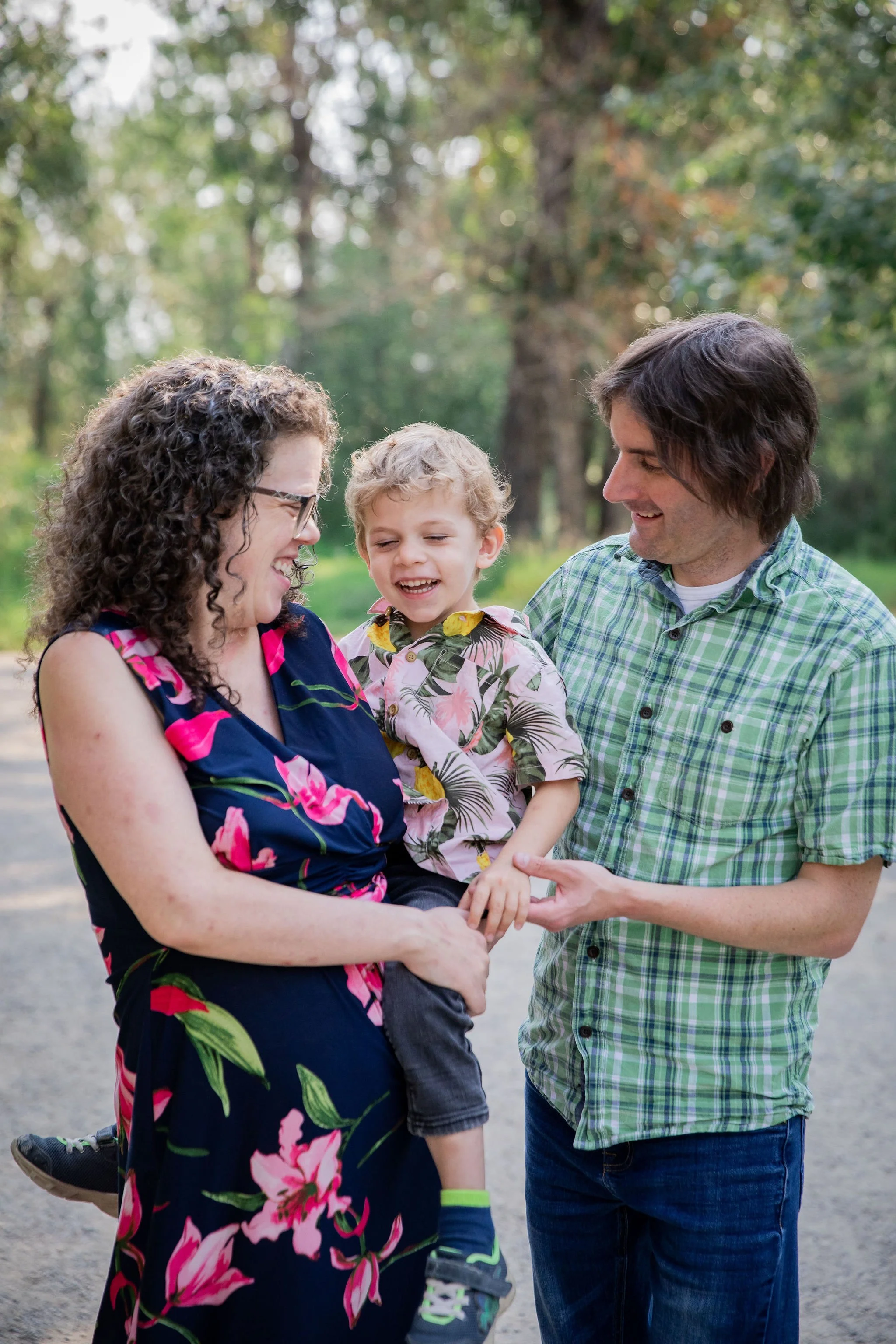 A family of three having fun outdoors, with the mother holding a young boy and the father touching his arm, all smiling and laughing in a park with trees in the background. maternity family photography at Bow Valley Habitat Station in Calgary Alberta