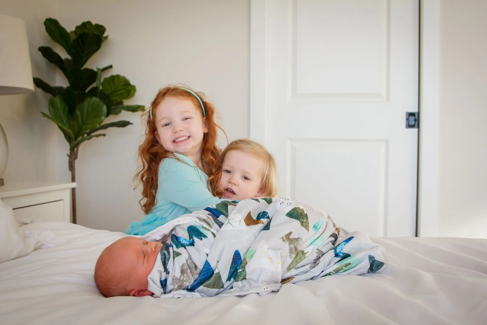 Two young girls hugging an infant on a bed in a bedroom, with a tall green plant and a white door in the background.