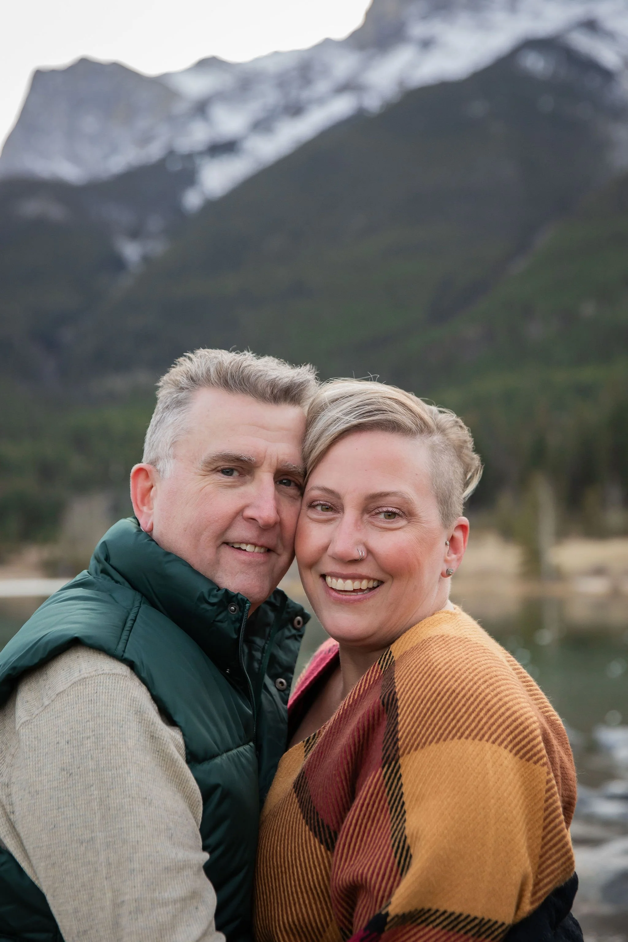 A smiling couple poses outdoors with a mountain landscape in the background, including snow-capped peaks, green hills, and a lake just a short drive from Calgary, Alberta 