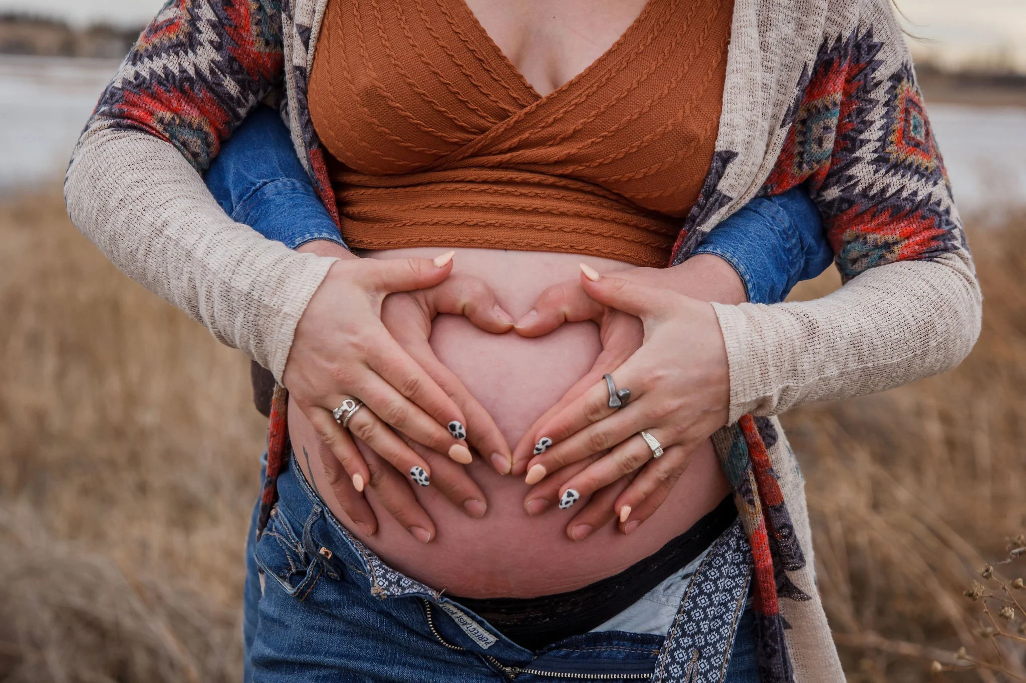 Close-up of a pregnant woman's belly being held by three pairs of hands, forming a heart shape, outdoors in a field. Maternity photography session by Ralph Klein park in Calgary, Alberta. 
