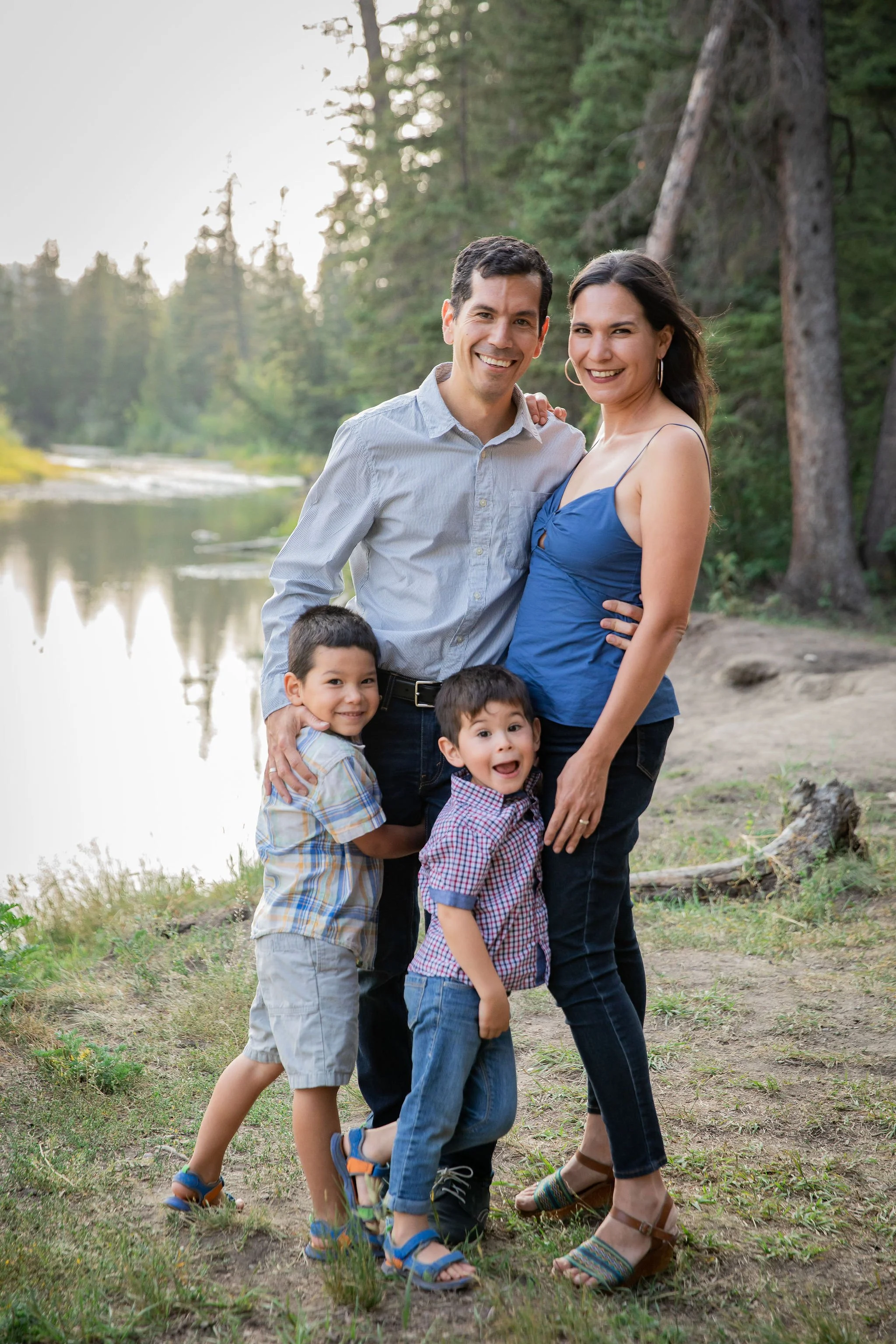 A happy family of four, two adults and two children, smiling outdoors near a lake with trees in the background. family phtography Bebo Grove, Calgary, Alberta. 