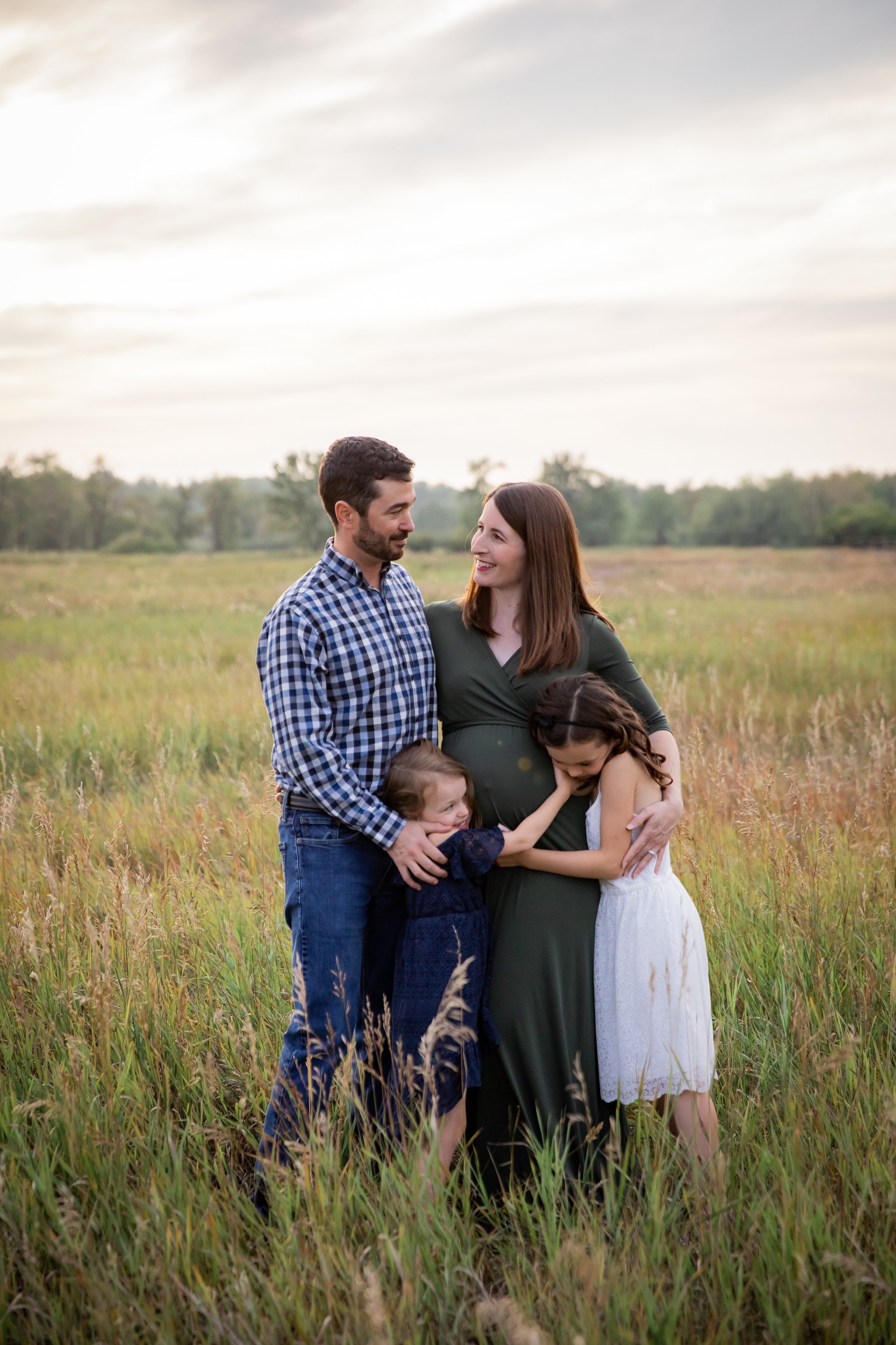 A family of four standing in a grassy field, with a man and woman embracing, and two young girls hugging them, during sunset. maternity session in Calgary, Alberta. 