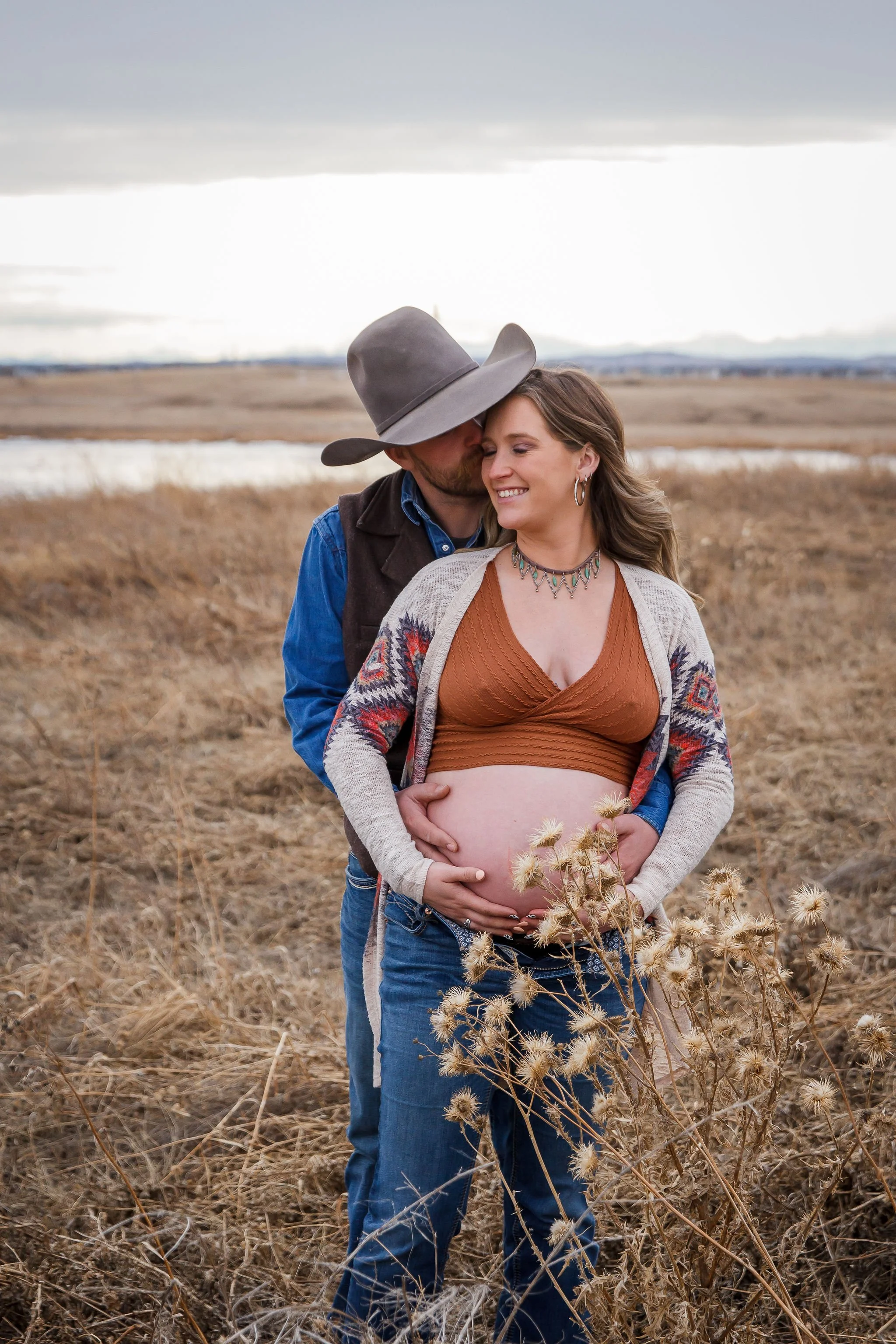 A pregnant woman smiling with her partner behind her in a field of dry grass, her partner kissing her forehead. The woman is cradling her belly, and both are holding her belly. Maternity photography session by Ralph Klein park in Calgary, Alberta. 