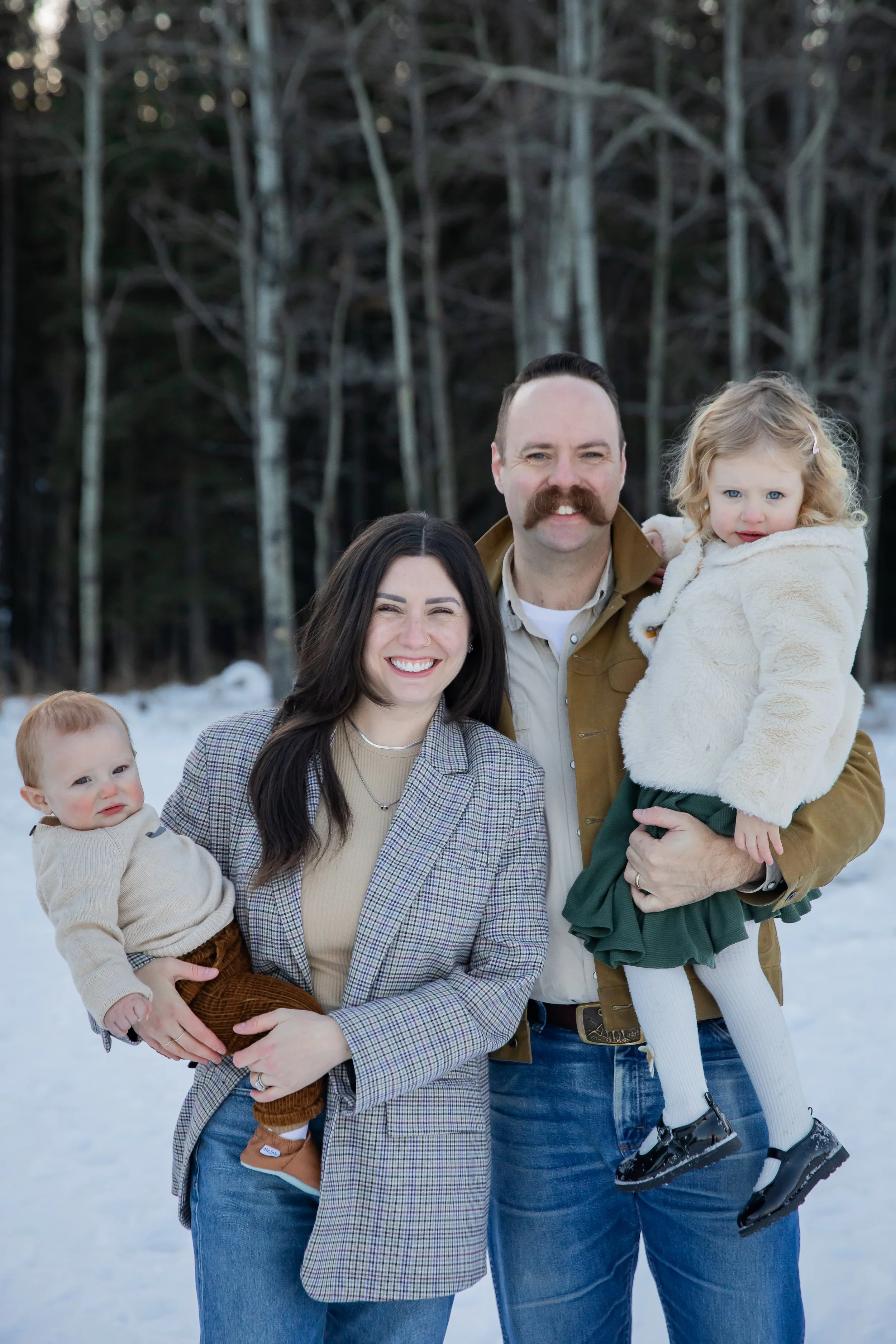 A family of four standing outdoors in snowy forest, smiling at the camera. The woman has long dark hair, the man has a mustache and is holding a young girl. The woman is holding a young boy, and the girl is wearing a white fluffy coat and black shoes