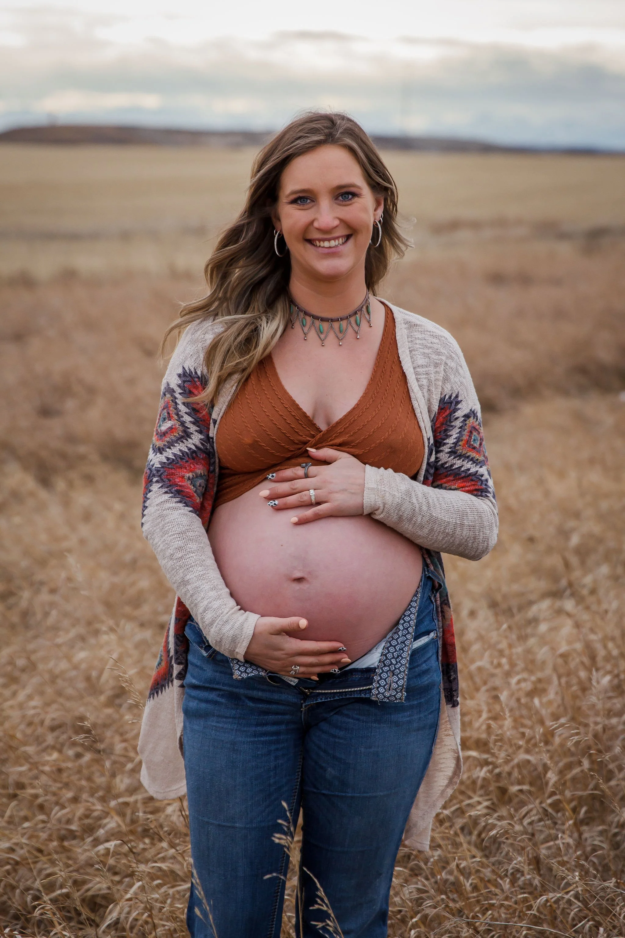 A pregnant woman standing in a field of dry grass, smiling and holding her belly with both hands, during a cloudy day. Maternity photography session by Ralph Klein park in Calgary, Alberta. 