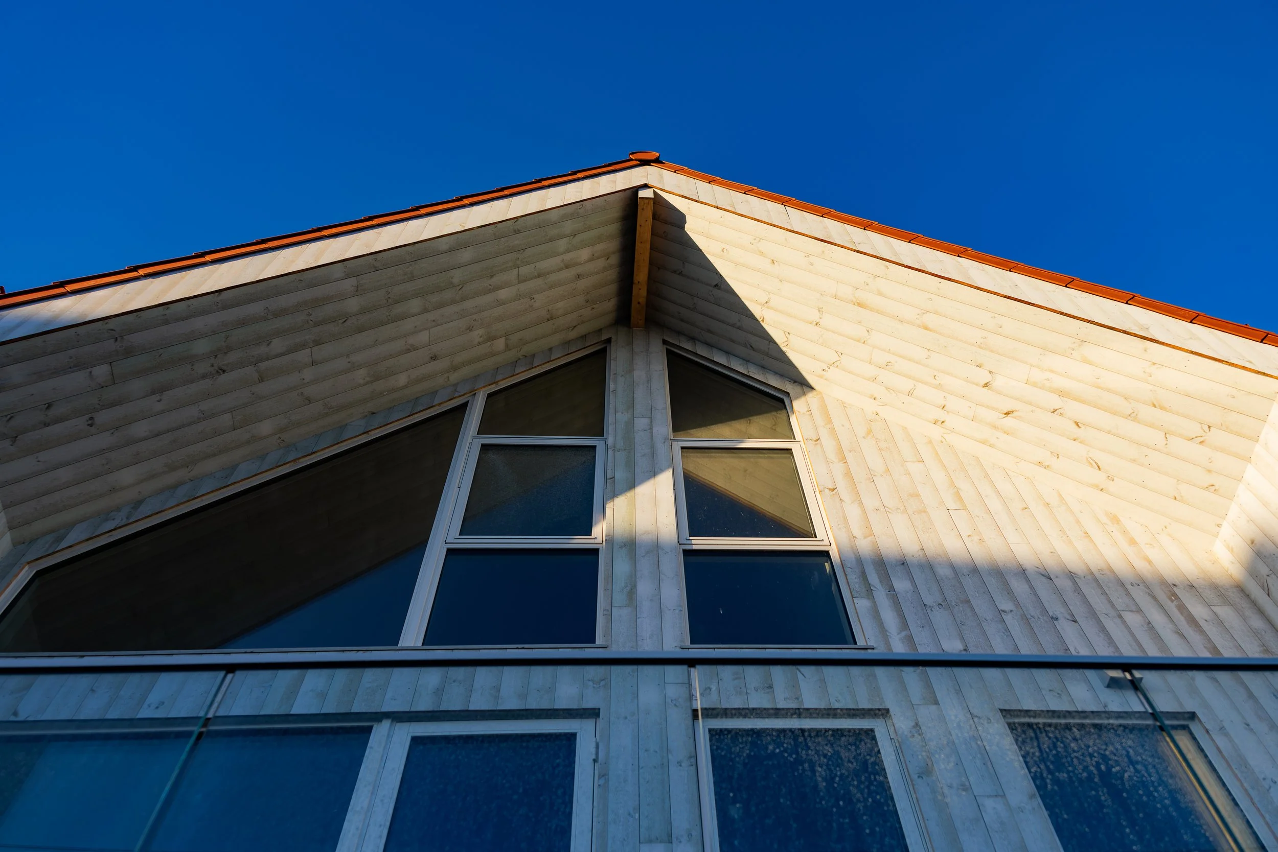 Nærbilde av et moderne hus på Risøy med trekledning og store vinduer, sett fra bunnen av, mot en klar blå himmel.