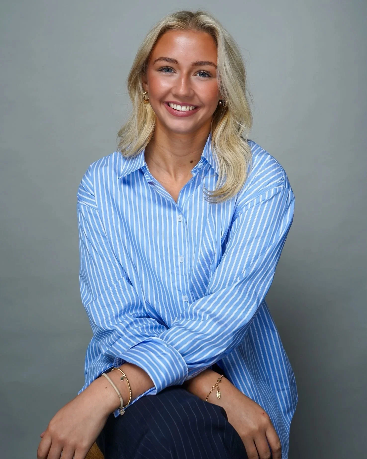 A smiling woman with blonde hair, wearing a blue and white striped shirt, sitting with her arms crossed and accessorized with gold jewelry, against a plain gray background.