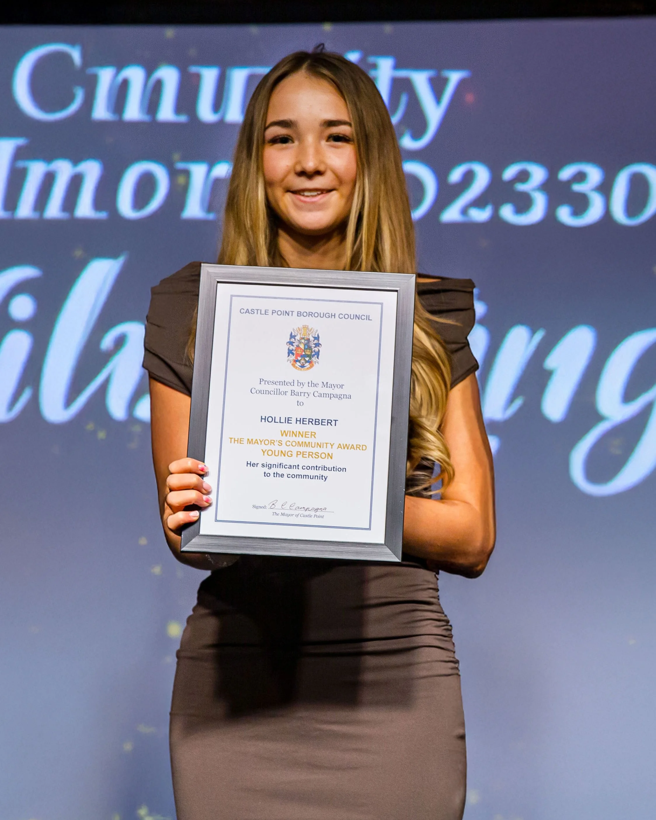 A young woman with long blonde hair smiling and holding a framed award certificate. She is wearing a brown dress with puffed sleeves. Behind her, there is a dark blue background with blue and white text, part of which says 'Courage' and some numbers.