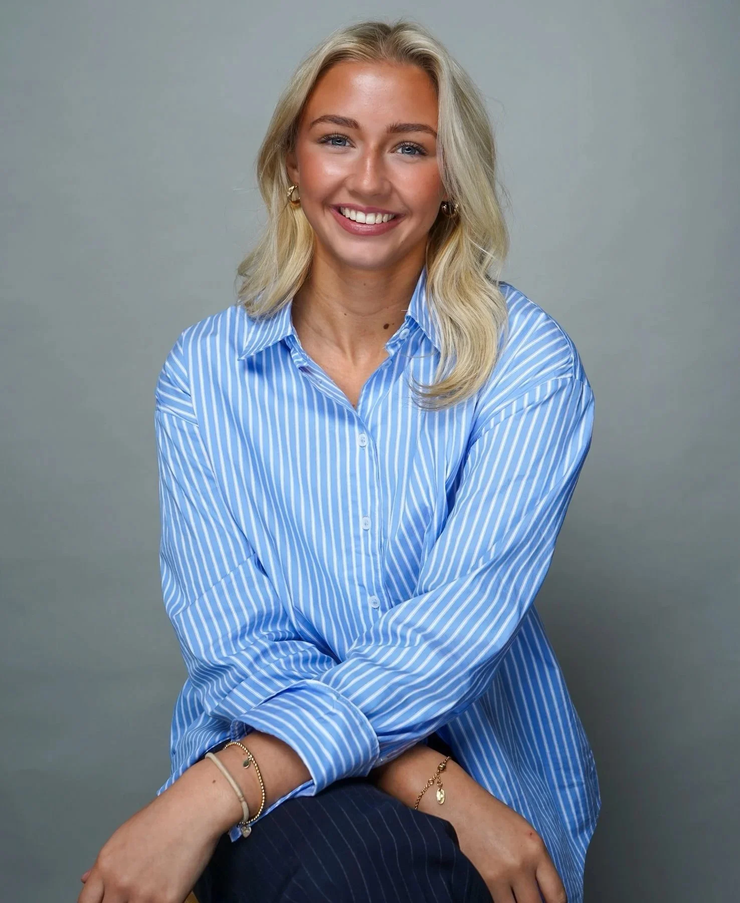 A woman with blonde hair, wearing a blue and white striped shirt, smiling, sitting against a plain gray background.