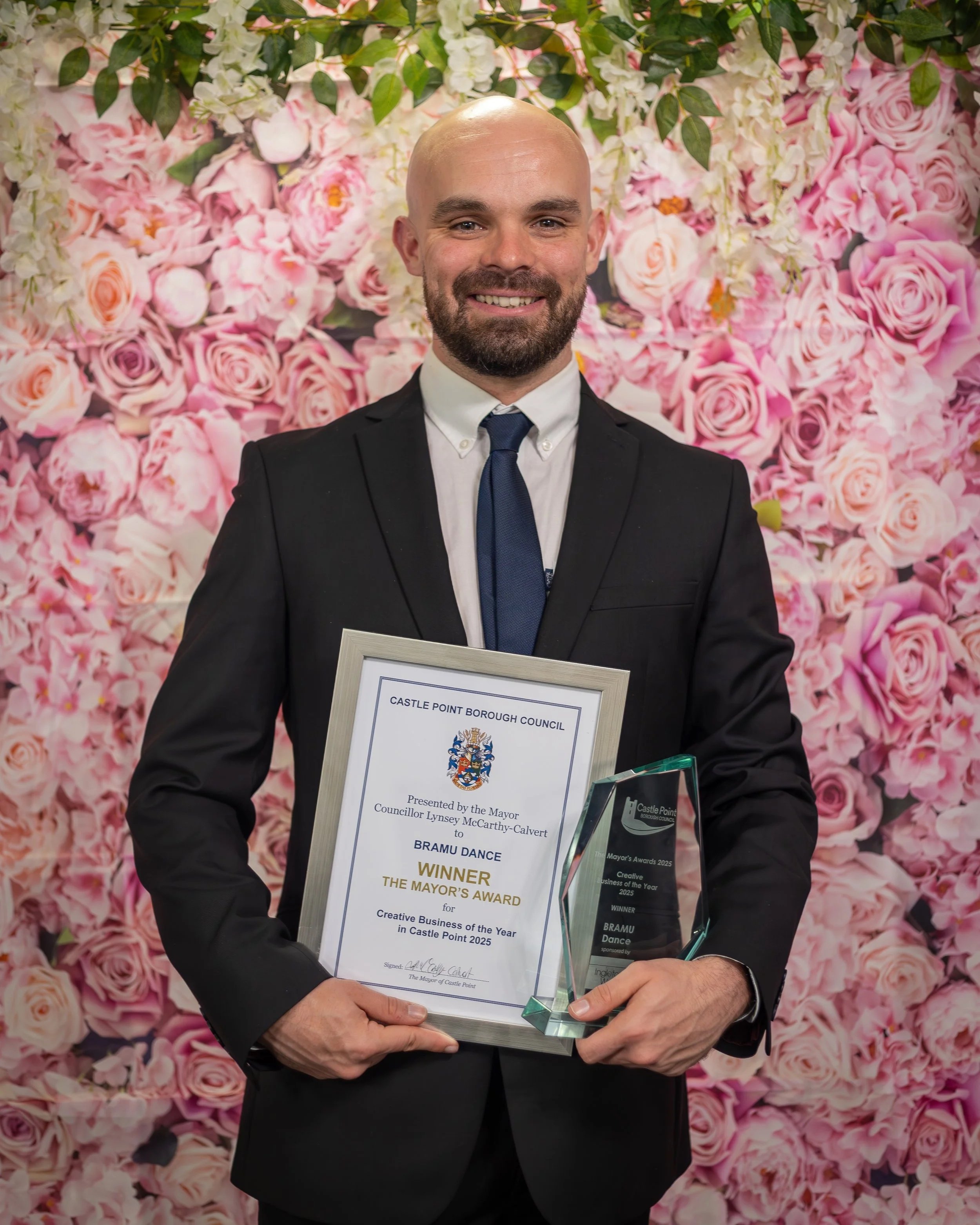 A man dressed in a black suit, white shirt, and dark blue tie, smiling and holding a framed certificate and a clear glass award. The background is a floral wall with pink and white roses and greenery.