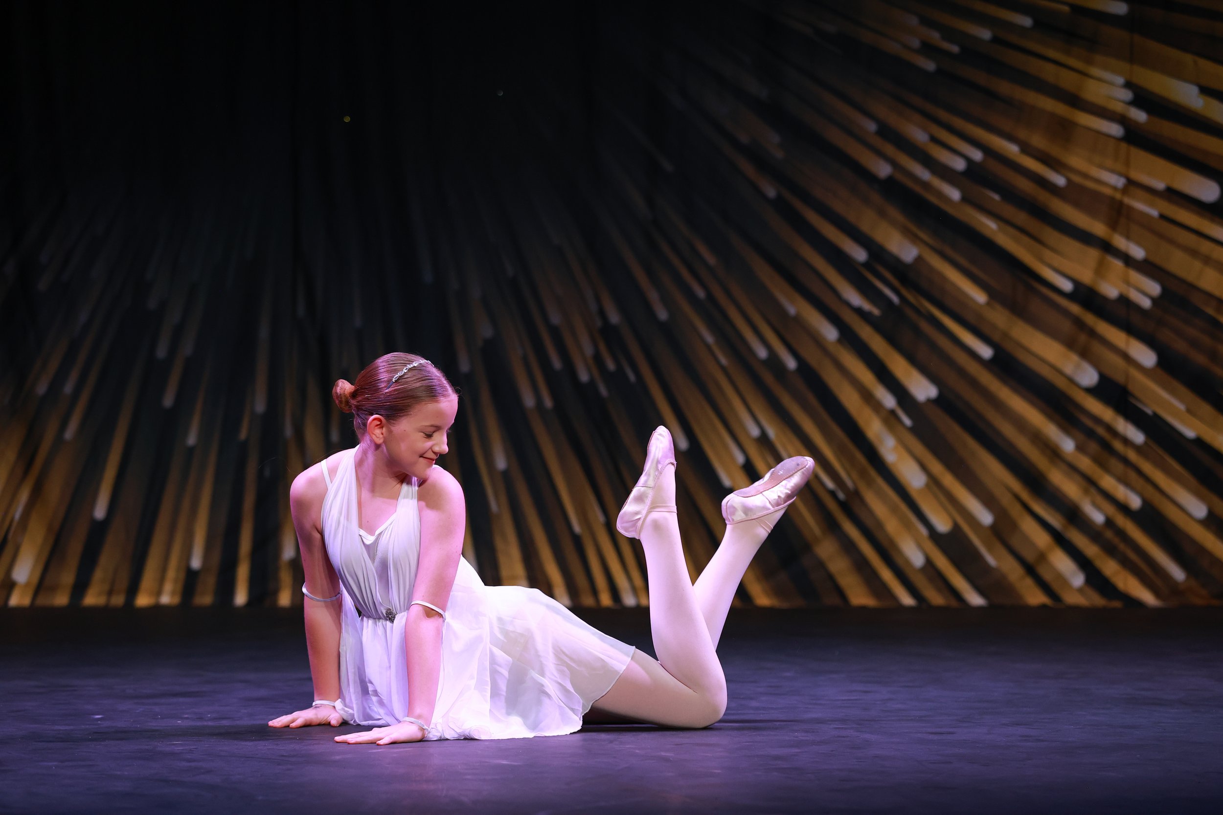 A ballerina in a white dress performing a pose on stage with a wooden stage backdrop.