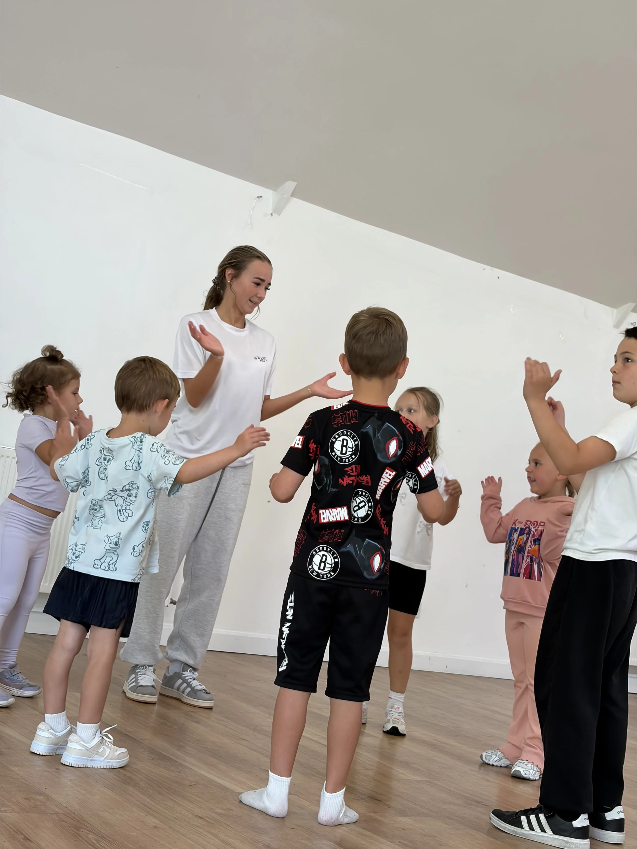 A group of children in casual clothing participating in a dance or exercise class led by a woman in a white t-shirt. They are inside a room with a wooden floor and white walls.