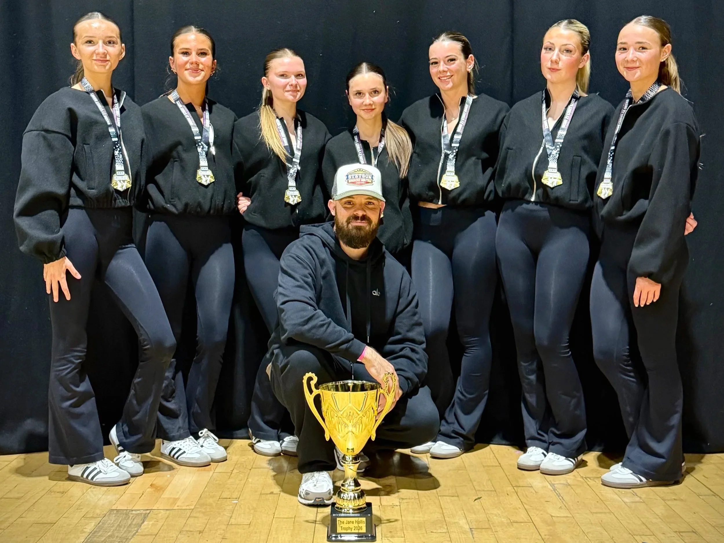 Group of seven women and one man posing with a trophy. The women are dressed in black sportswear and the man is in black hoodie and cap. The trophy has a plaque that reads 'The Jane Hollis Trophy 2020'.