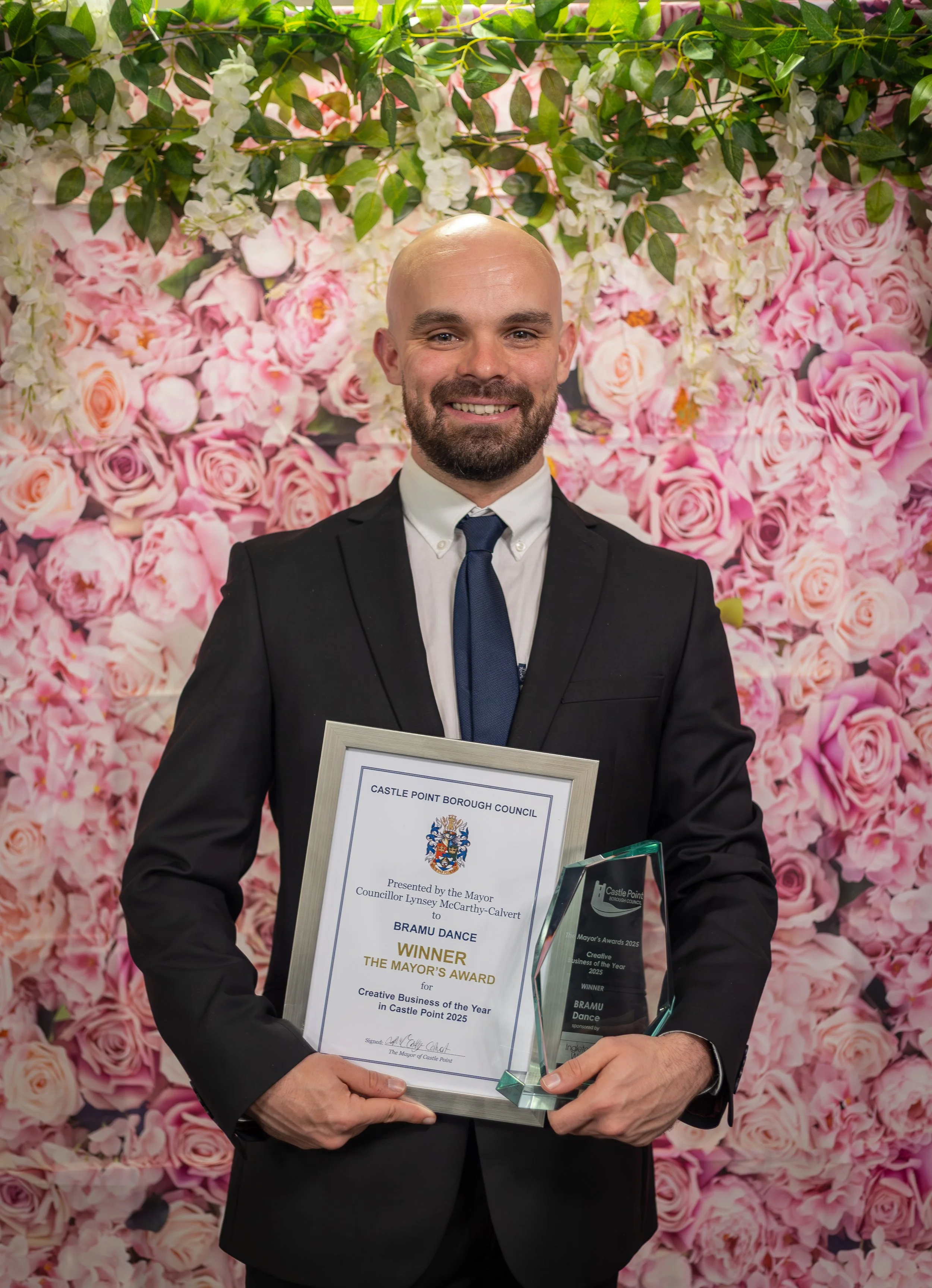 A man in a black suit, white shirt, and navy tie holding an award and certificate, smiling in front of a pink and white rose floral backdrop.