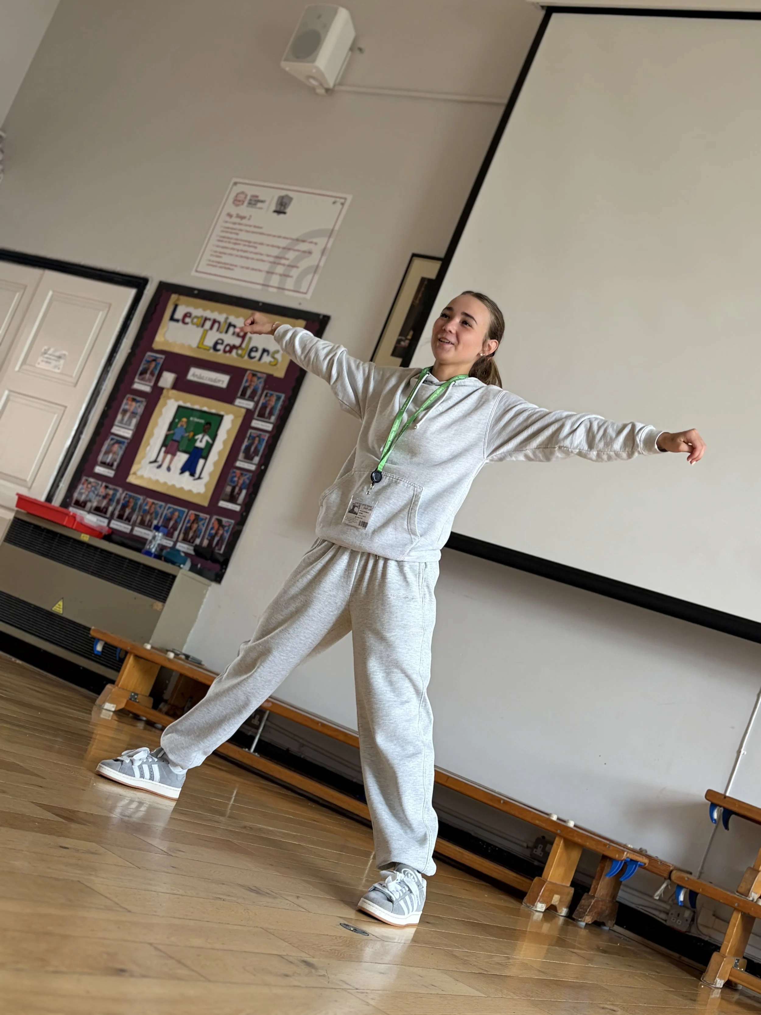 A young woman with long brown hair tied back, wearing a light gray hoodie, matching sweatpants, and gray and white Adidas sneakers, standing with arms outstretched in a classroom or gymnasium.