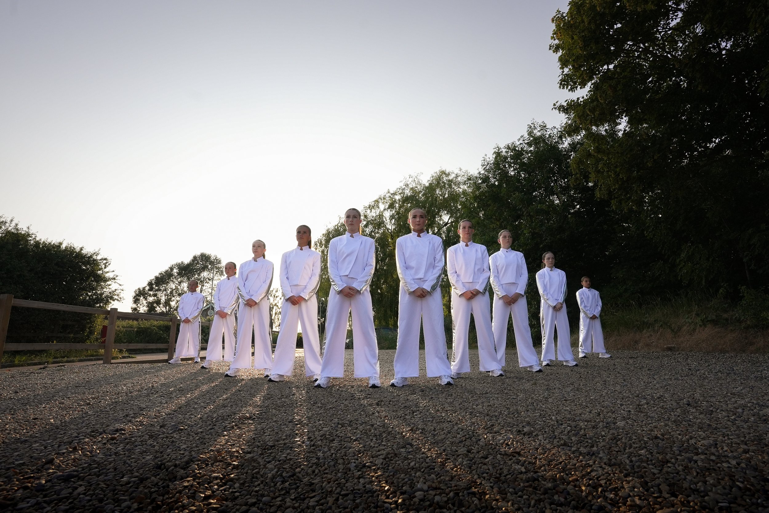 Group of women standing outdoors in white uniforms during sunset, with trees in the background.