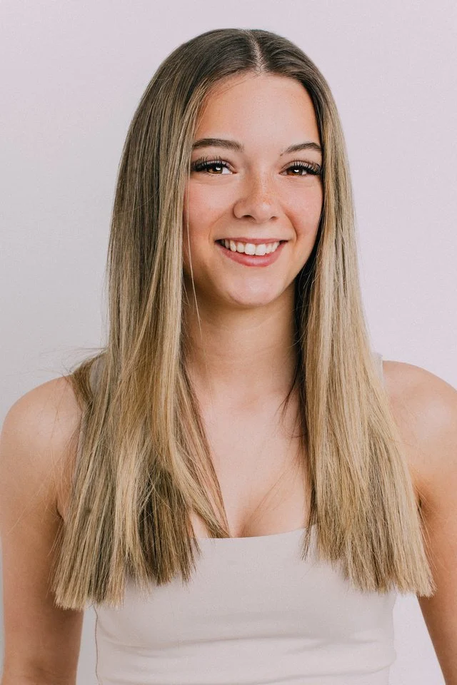 Young woman with long straight blonde hair smiling at the camera, wearing a white sleeveless top against a plain light background.