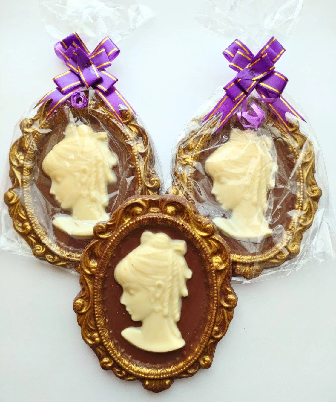 3 cameos made from chocolate on a white background. 2 are in cellophane bags tied with purple bows.