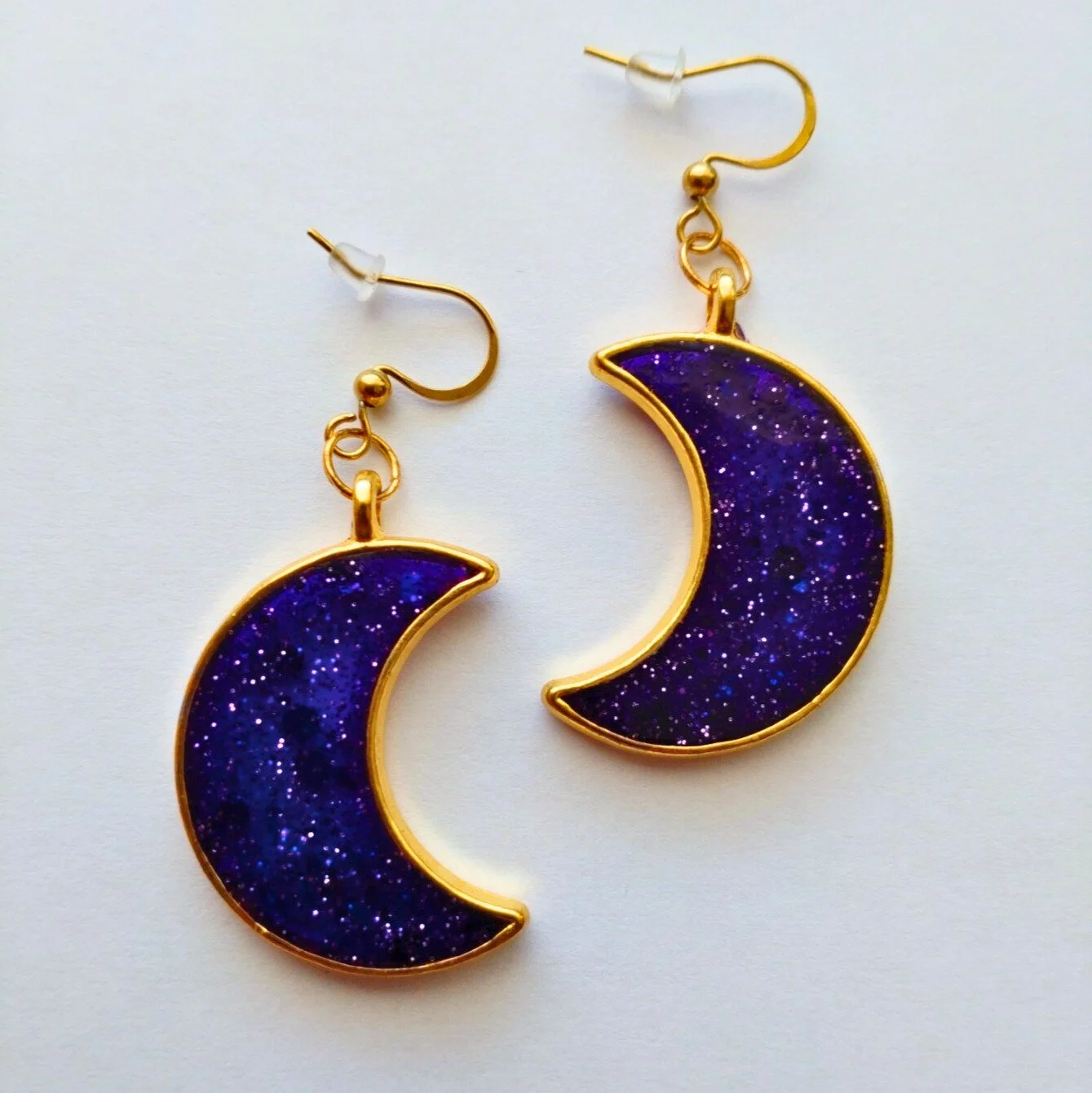 Gold coloured crescent moon earrings in glittery purple resin on white background