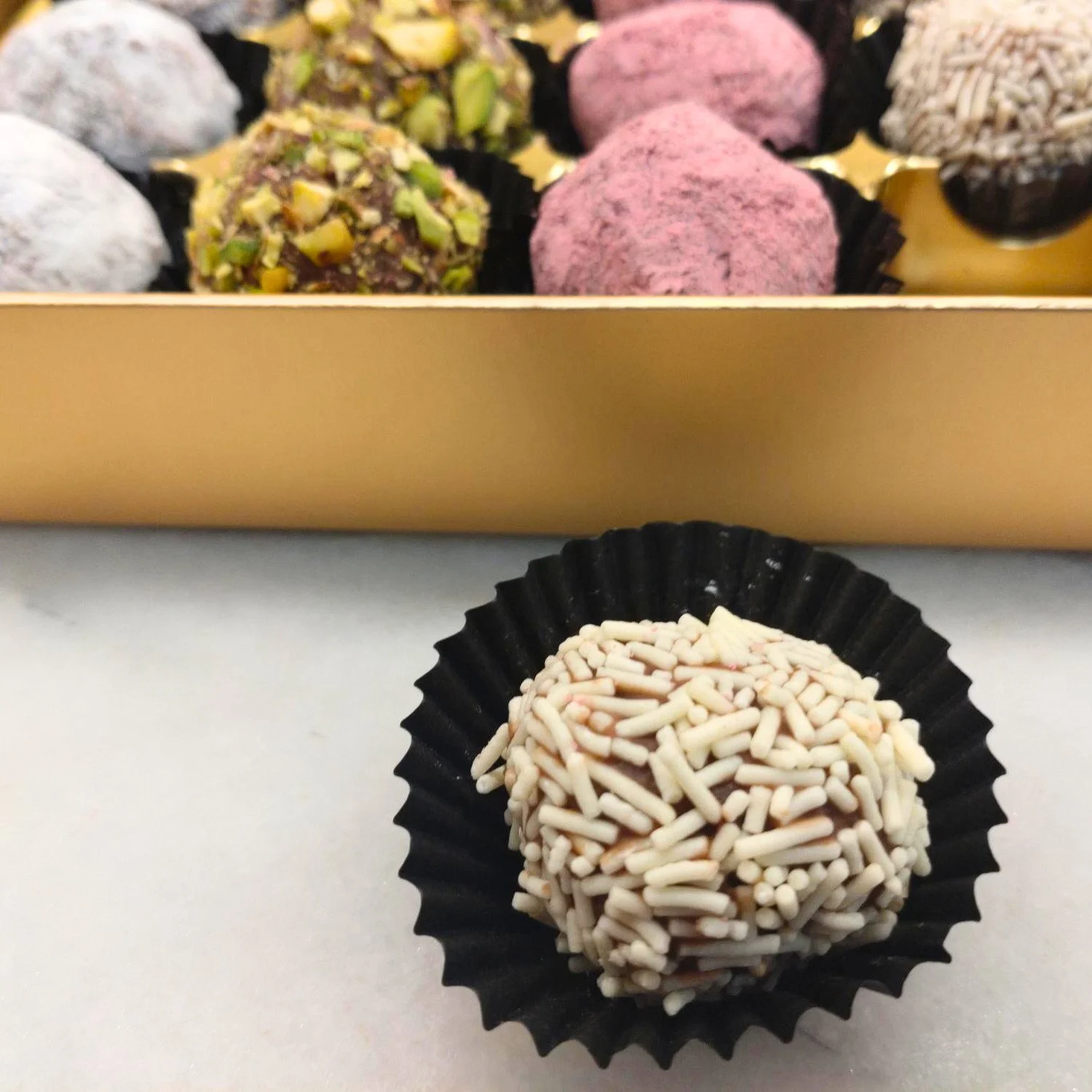 Close-up of a handmade milk chocolate truffle coated in white chocolate vermicelli, with assorted truffles in the background.