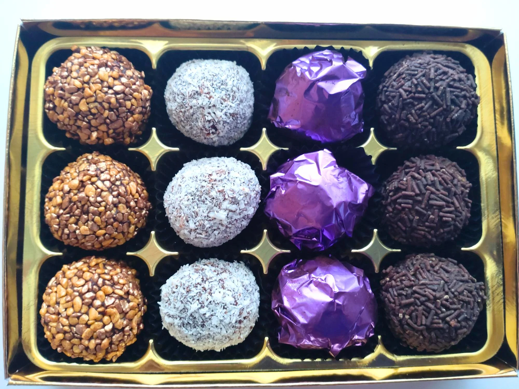 Overhead view of 12 handmade dark chocolate liqueur truffles in four varieties, with bronze crunch, coconut, purple foil and chocolate vermicelli coatings.