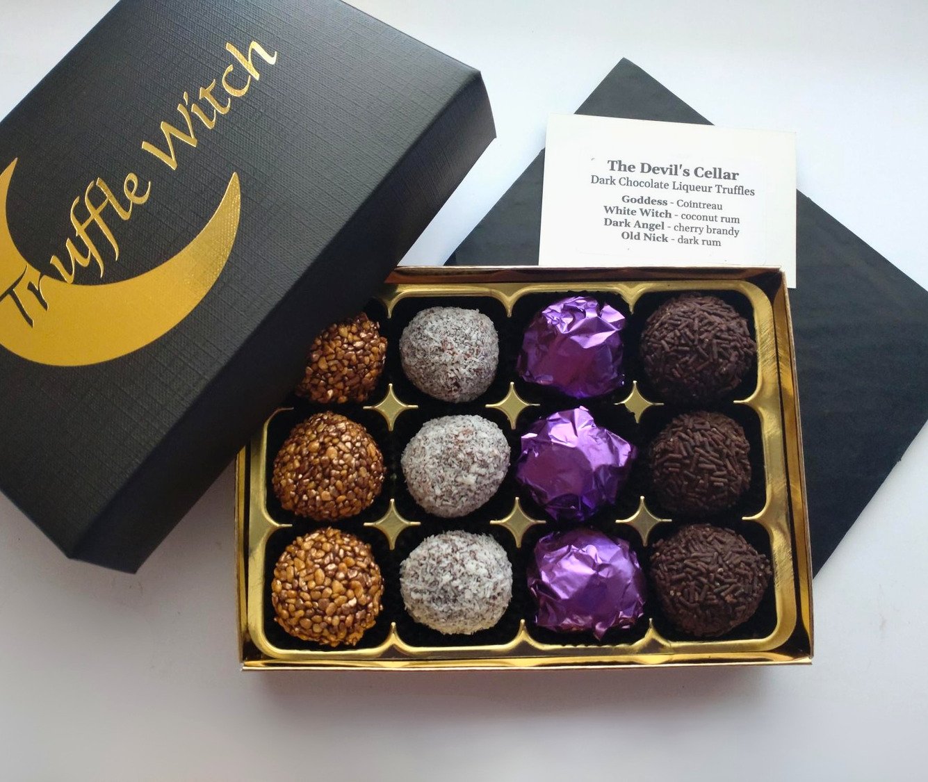 Luxury black Truffle Witch gift box with gold crescent moon logo, partially opened to reveal 12 handmade dark chocolate liqueur truffles and a printed flavour guide.