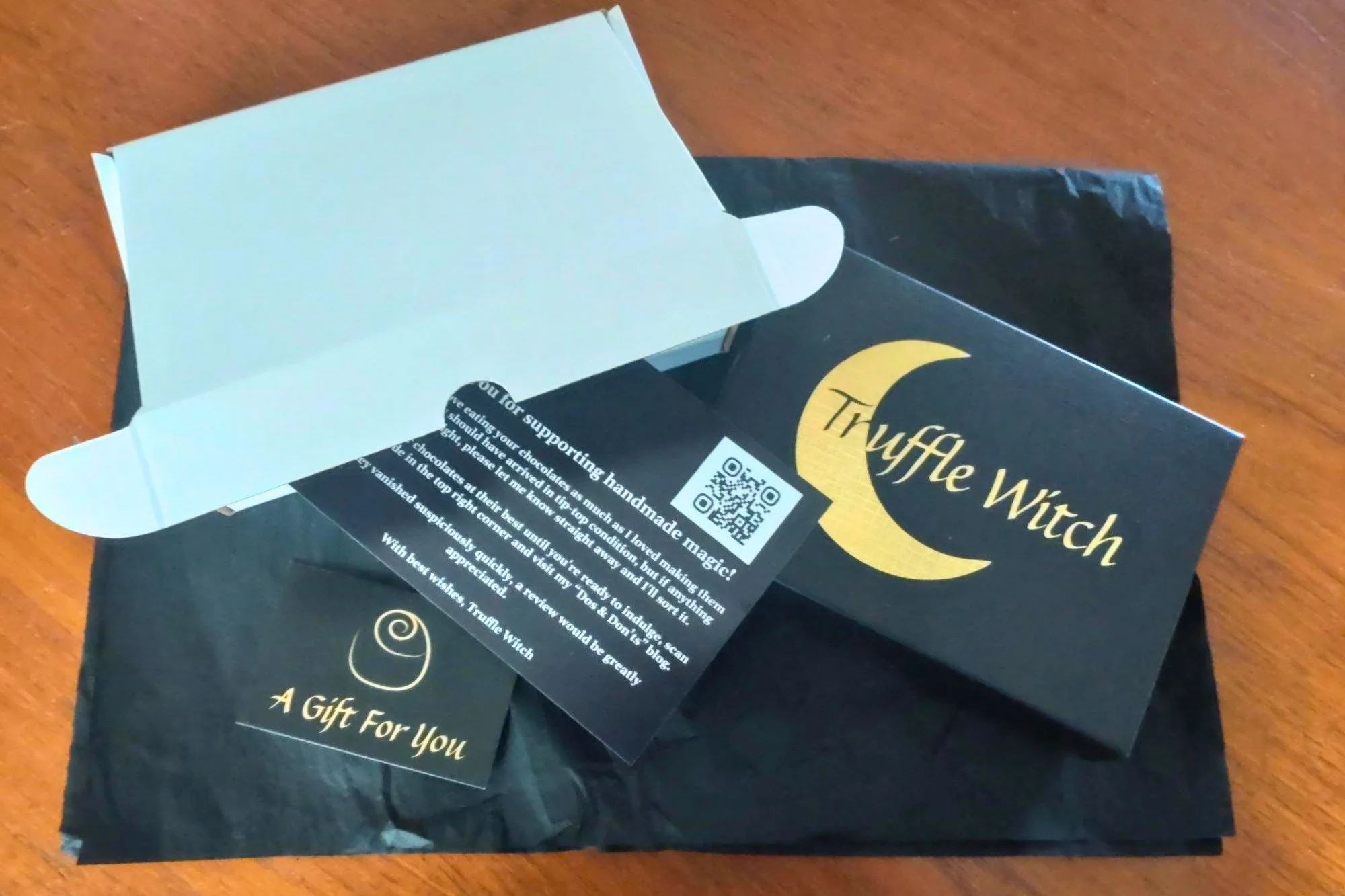 A white postal box lying on black tissue paper beside a black chocolate box with gold Truffle Witch logo. A printed flyer and gift card are also on the tissue paper