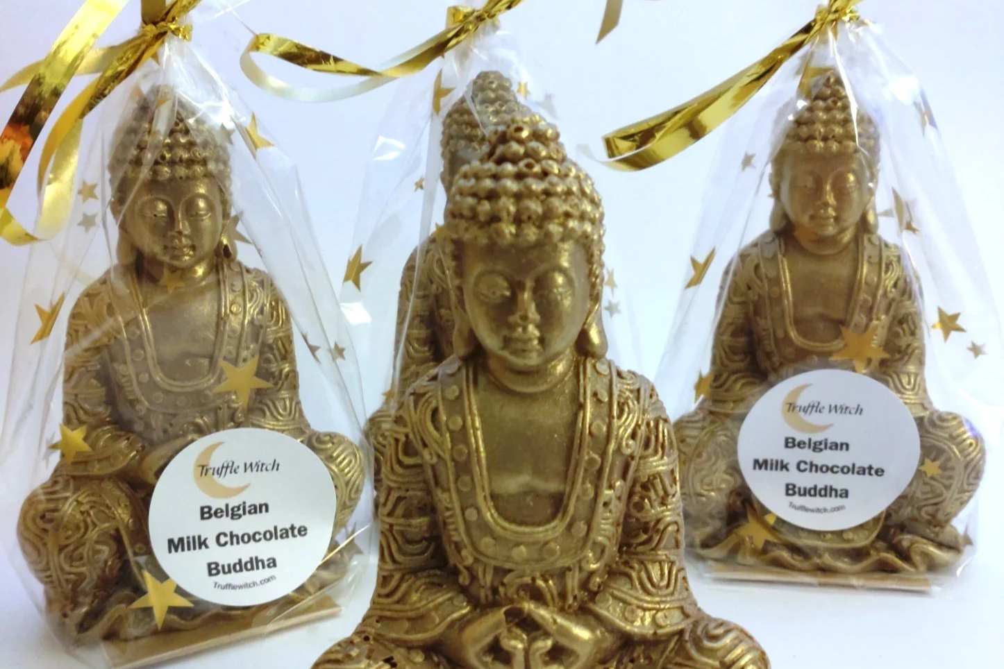 Photograph shows 4 Buddha figures made from chocolate. They are dusted with gold lustre and 3 are in cellophane bags decorated with gold stars and tied with gold ribbon