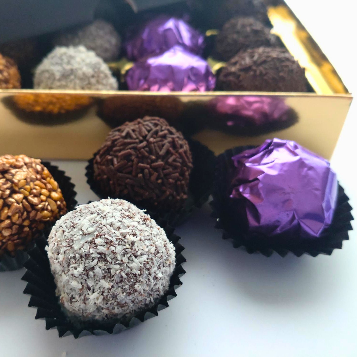 Close-up of four handmade truffles featuring bronze sugar crunch, coconut, dark chocolate sprinkles and a purple foil-wrapped chocolate, displayed in black paper cases.
