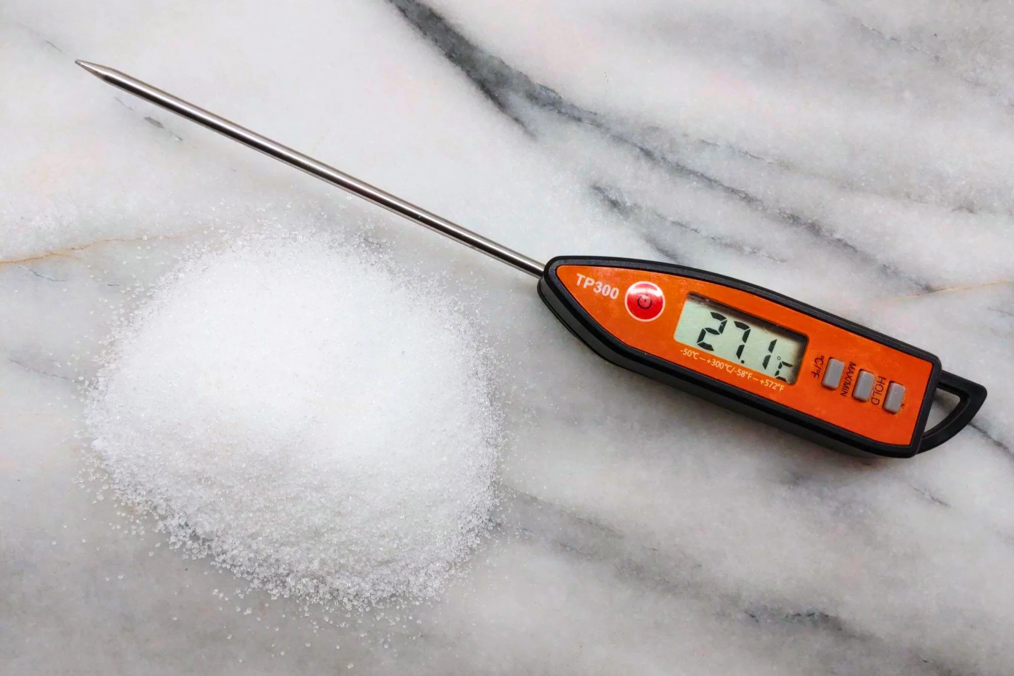 An orange and black digital thermometer reading 27.1 degrees C on a grey marble background beside a small pile of white sugar